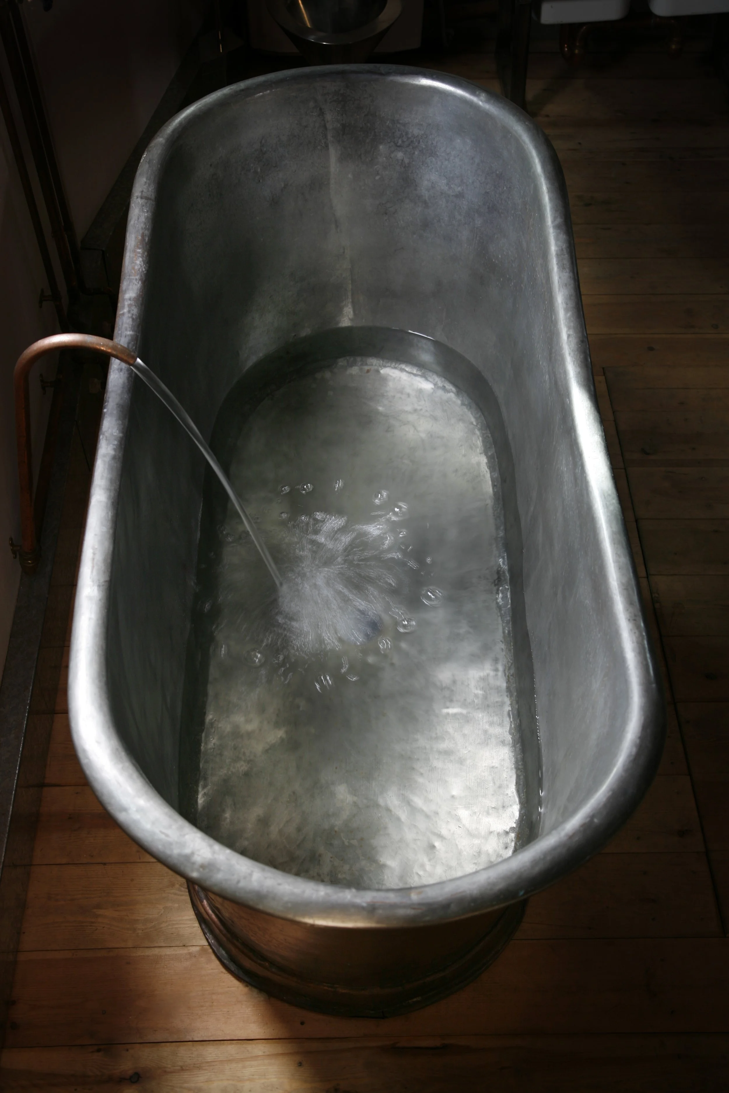 A vintage metal bathtub with water flowing from a copper pipe, situated on a wooden floor.