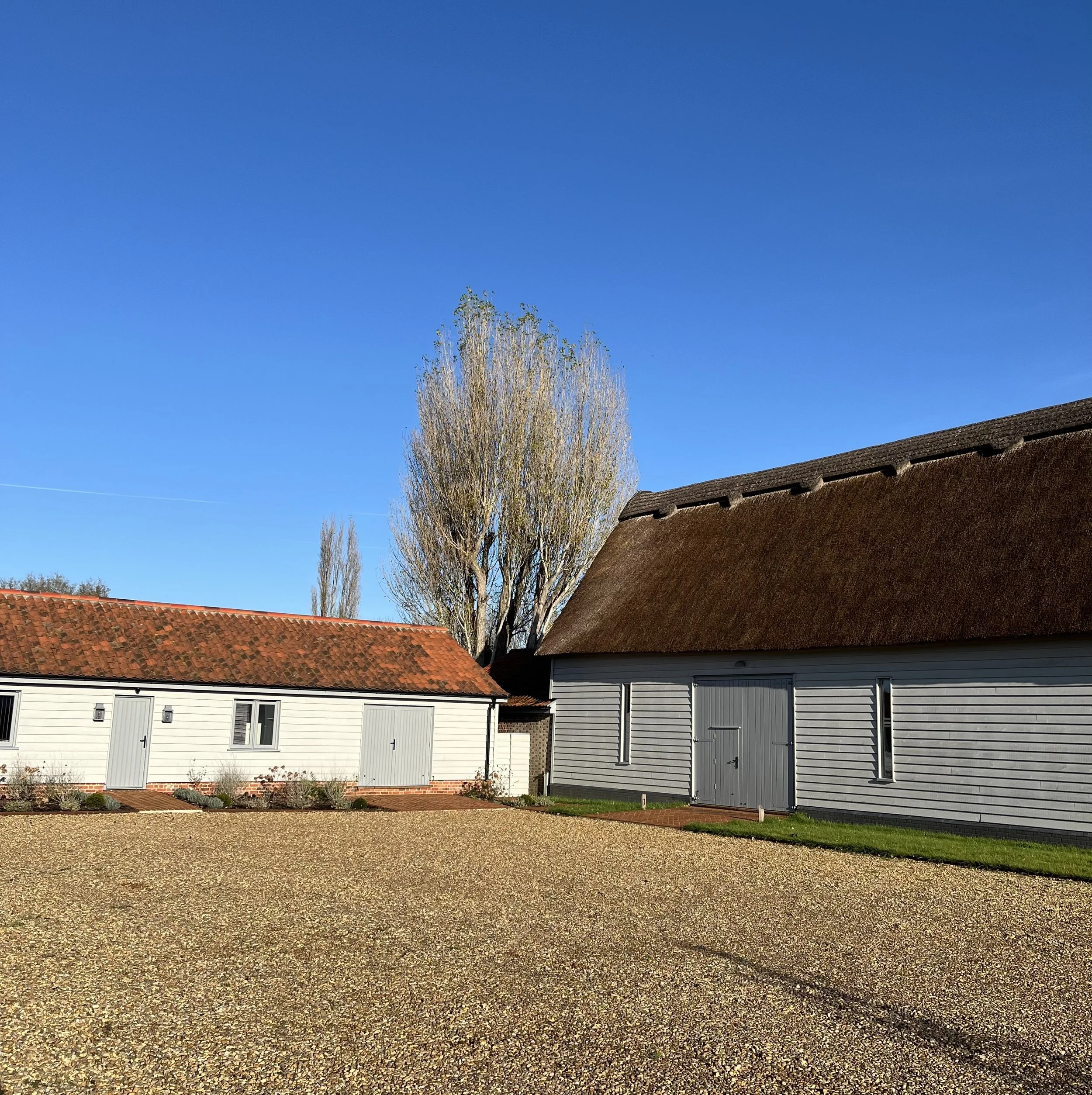 Two farm buildings with gravel yard, one with white siding and red tile roof, the other with grey siding and brown roof, under clear blue sky, with tall tree in background.