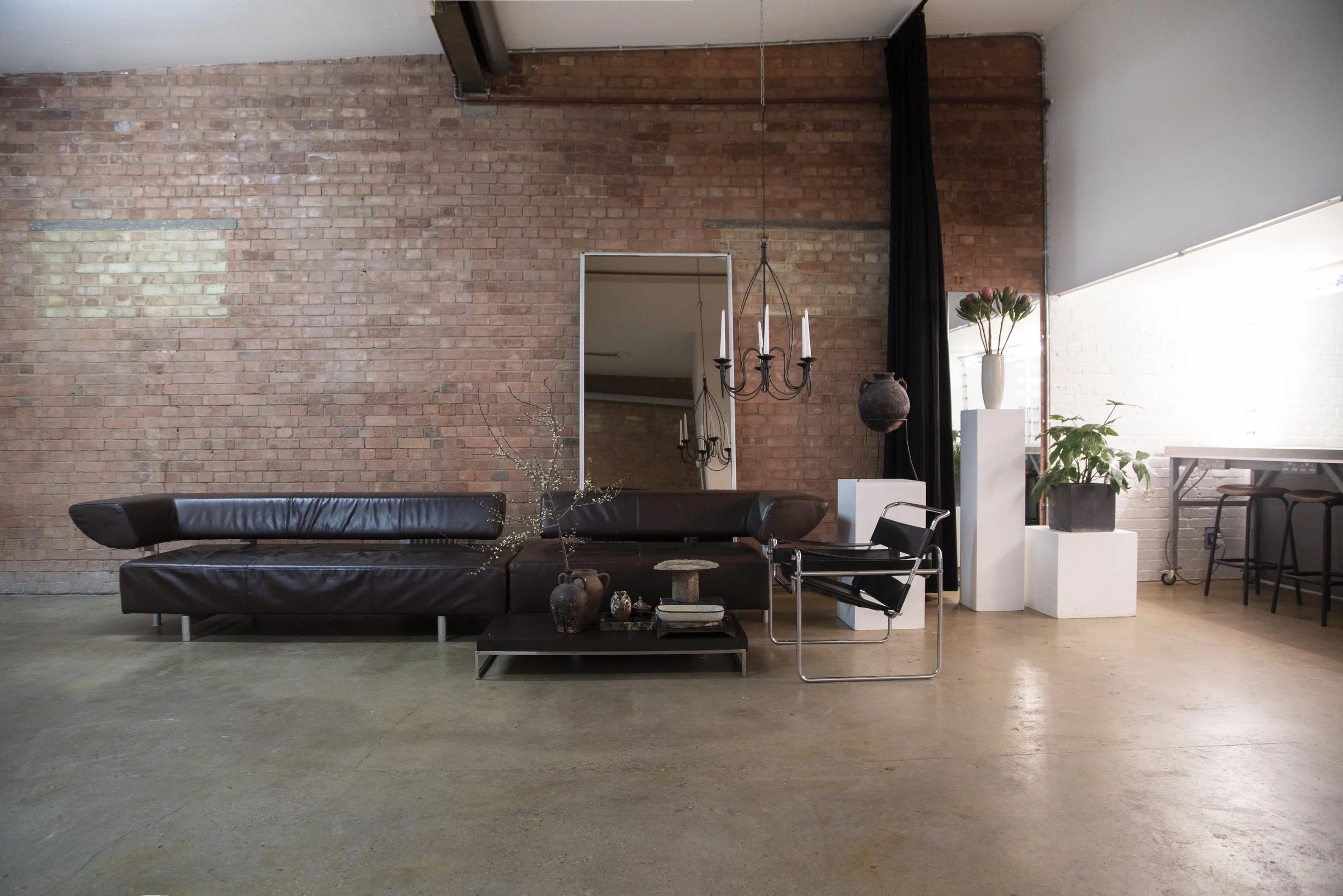 Modern living room with a brick wall, black leather sofa, various plants, white pedestals, a mirror, and a black chandelier.