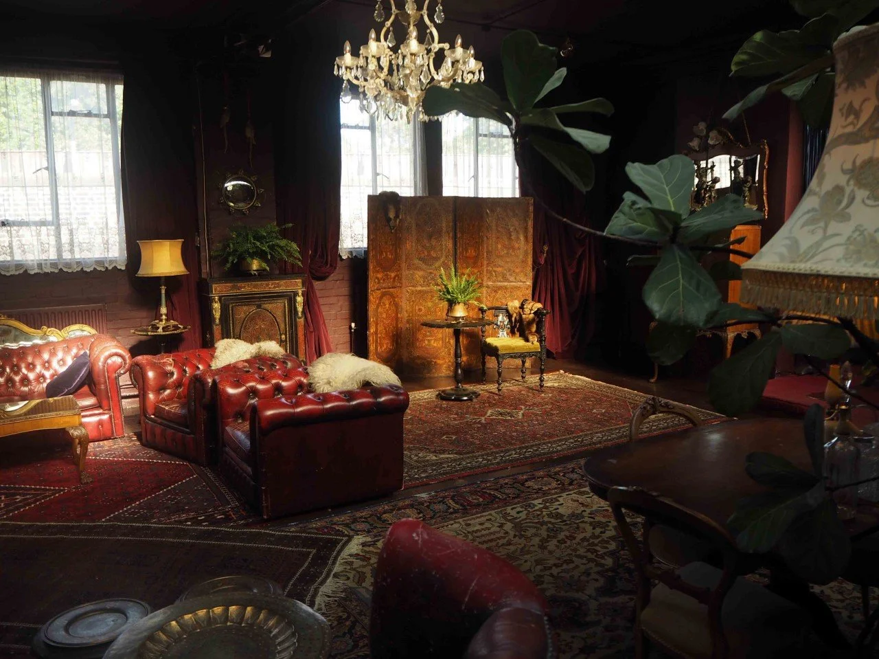 Vintage living room with red leather Chesterfield sofas, a chandelier, ornate furniture, and large windows with lace curtains.