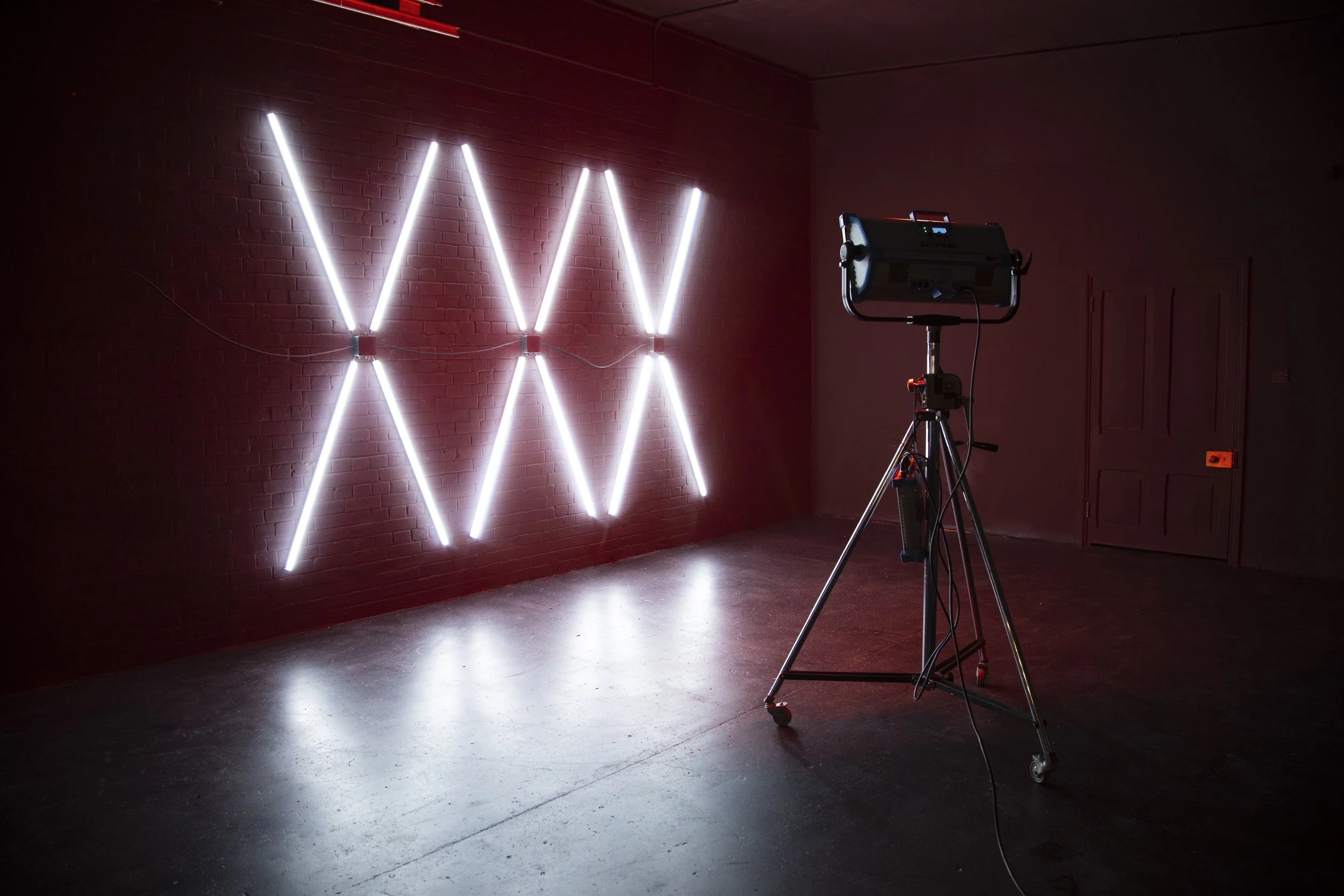 An art installation with vertical and diagonal white neon lights forming a pattern on a dark red brick wall, with a spotlight on a tripod in a black room.