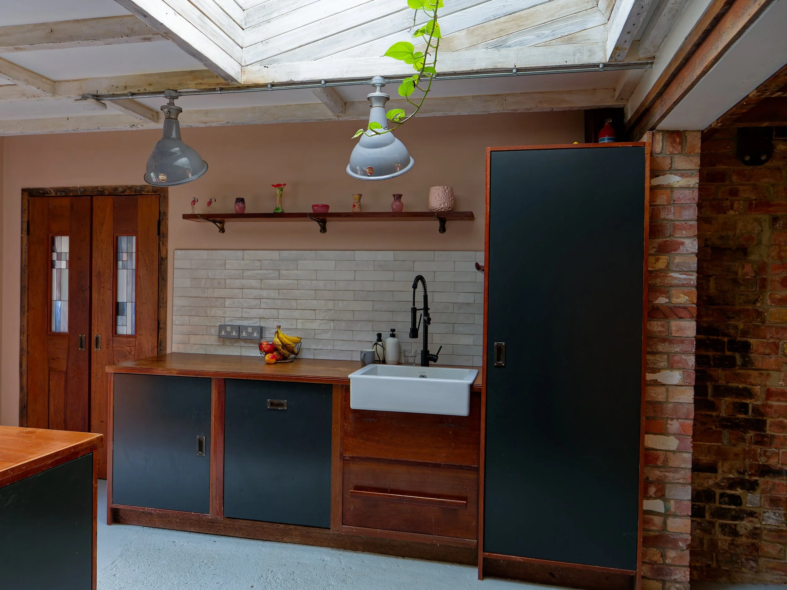 Kitchen with wooden cabinets, white brick backsplash, hanging shelves with decorative items, black faucet, white sink, fruit bowl on the counter, and exposed brick wall on the right.