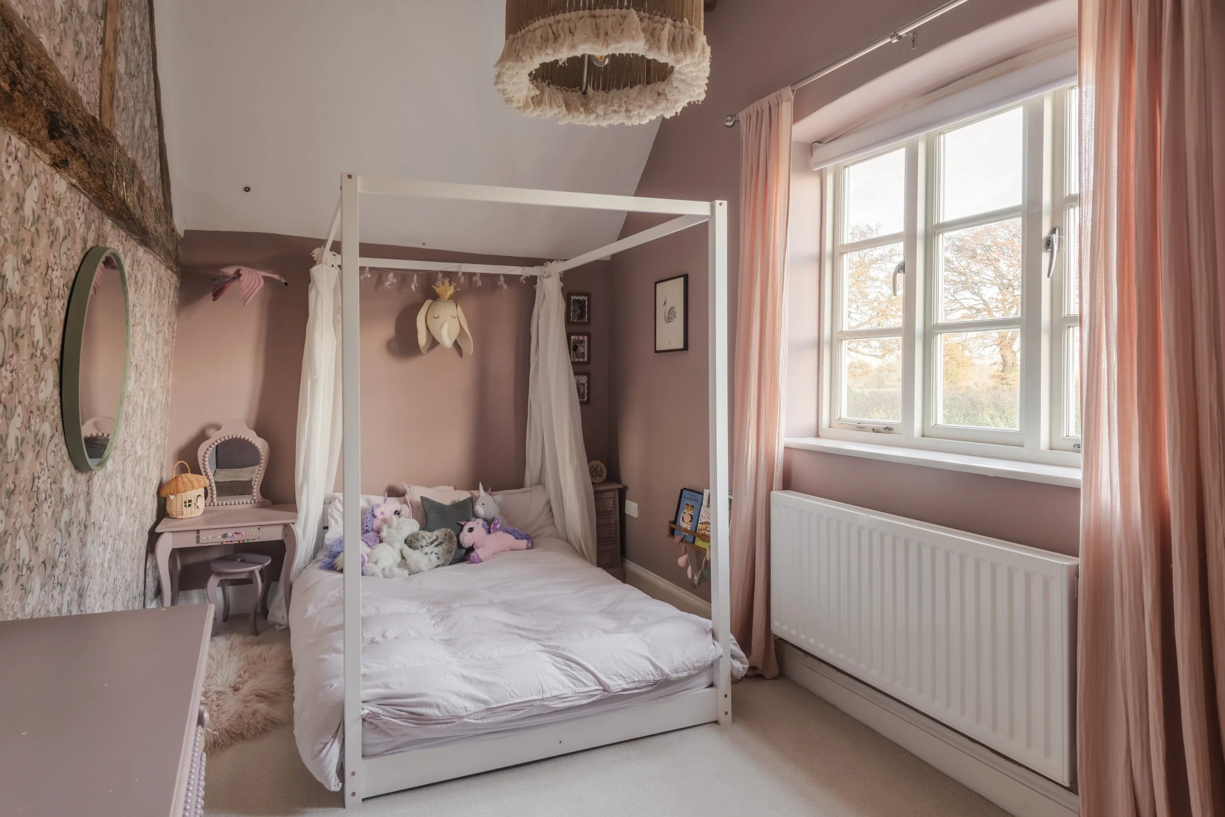 A cozy bedroom with a white canopy bed decorated with plush stuffed animals and unicorns. There are pink curtains and natural light coming through a large window. The room features soft, pastel colors and whimsical decor.