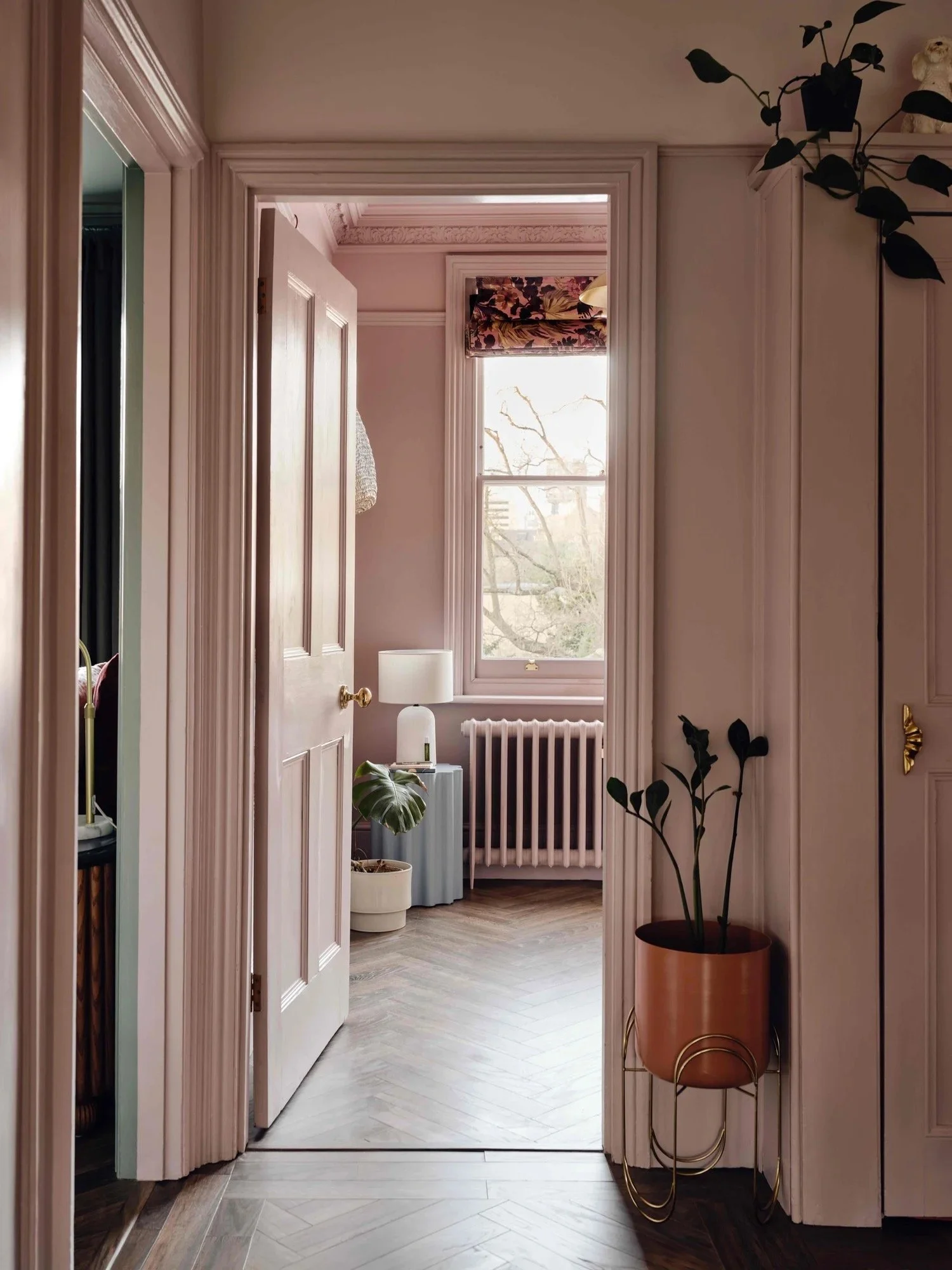 View of a cozy, well-lit room through an open door with a window showing trees outside. The room has a radiator, a small table with a white lamp, and potted plants, including a large-leafed plant near the window and another on a stand near the wall.