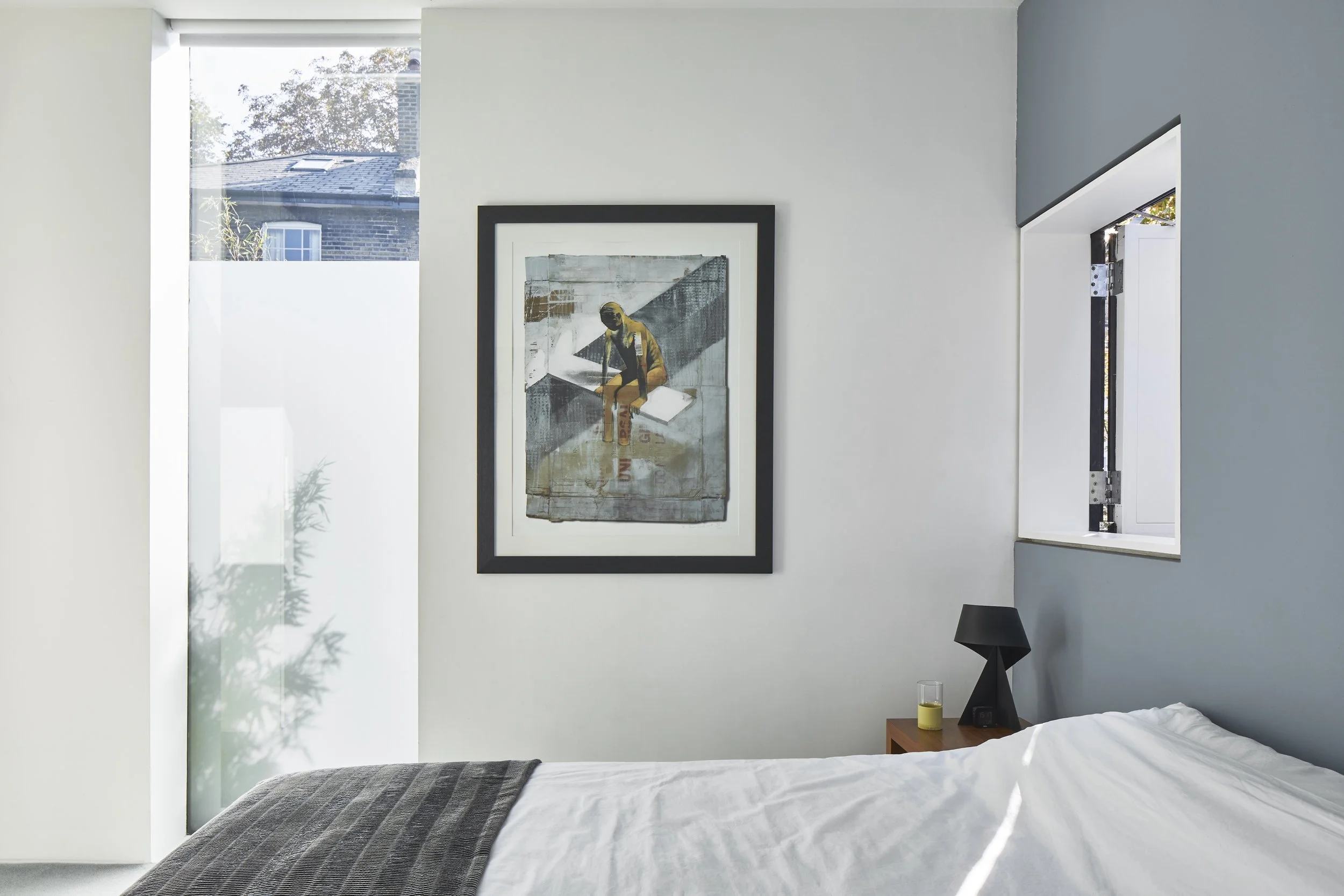 Modern bedroom with white and gray walls, a bed with white sheets and gray blanket, a bedside table with a black lamp and a glass of water, a large framed artwork on the wall, a window with open shutters, and sunlight streaming in.