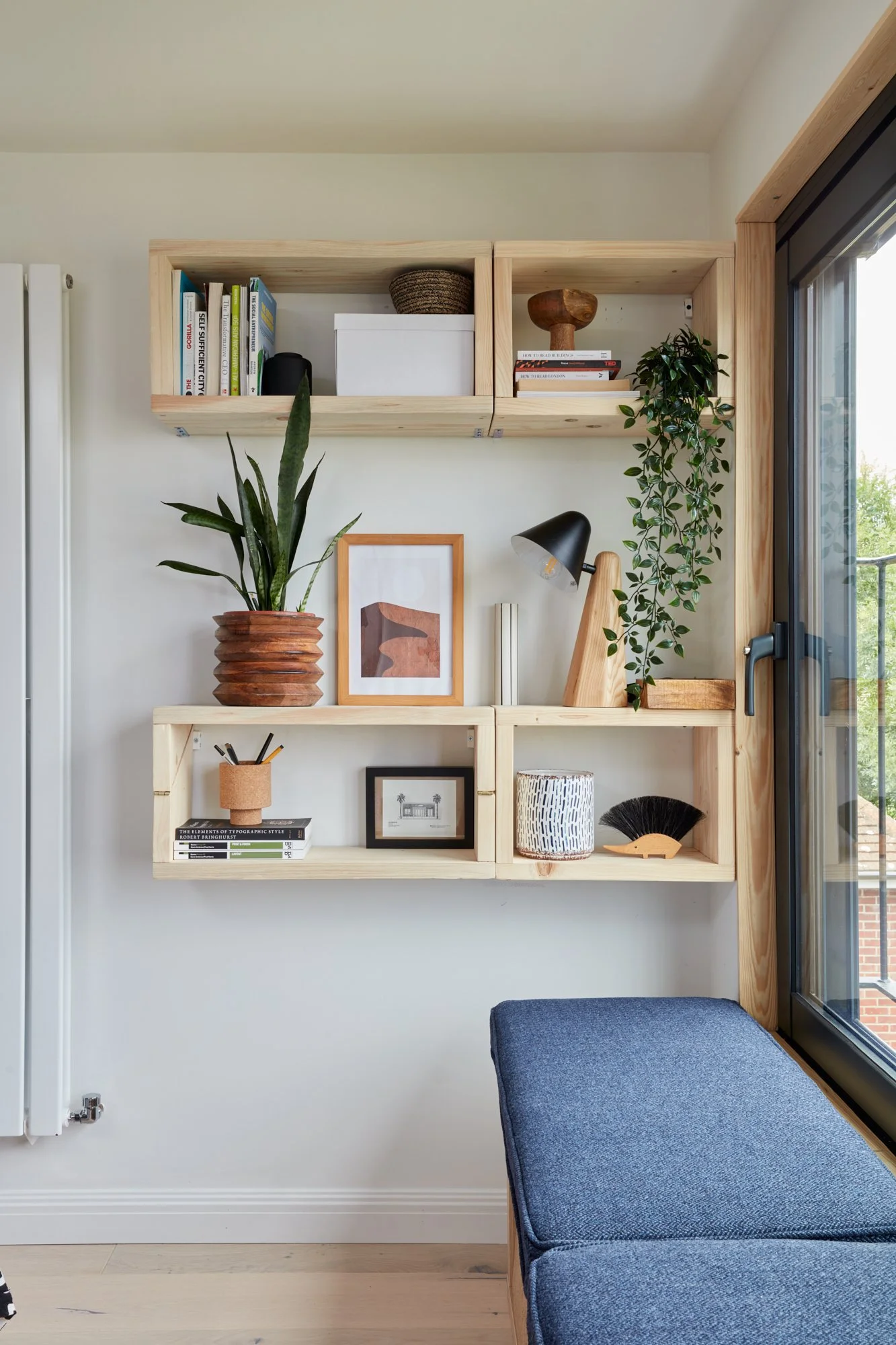 Decorative shelving unit with books, plants, picture frames, and decorative objects, next to a window and a blue upholstered seat.