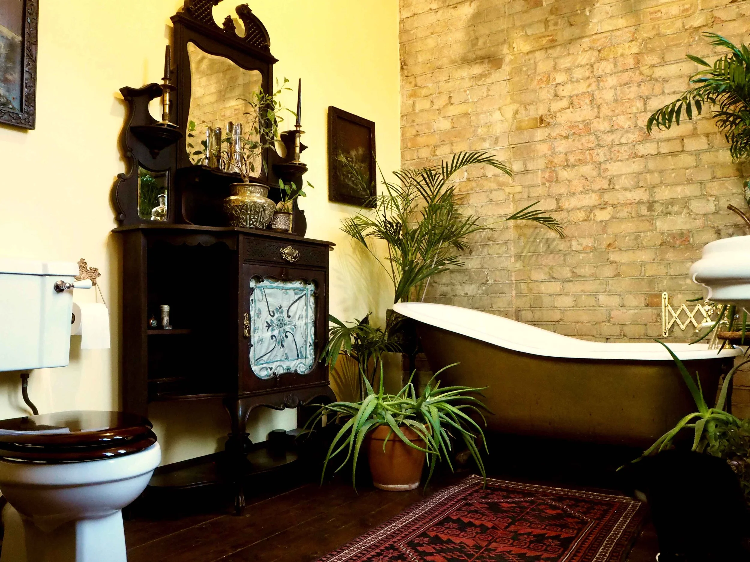 Vintage bathroom with a clawfoot bathtub, antique wooden cabinet, potted plants, brick and painted walls, and a decorative rug.