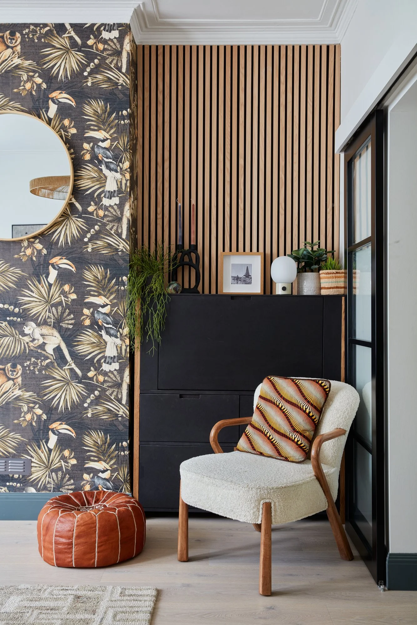 A corner of a modern living room with a beige armchair, a leather pouf, a black cabinet, a mirror, decorative objects, and a floral wallpaper with toucans and palm leaves.