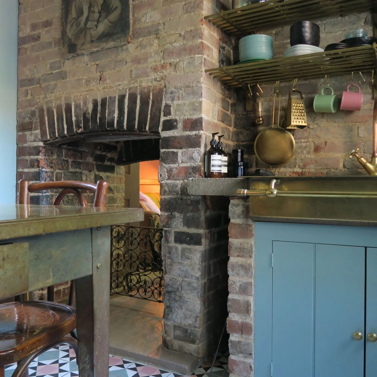 Kitchen brick wall with open brick fireplace, open shelves with dishes and cups, and a blue cabinet with brass sink and faucet. A table with wooden chairs is in the foreground.