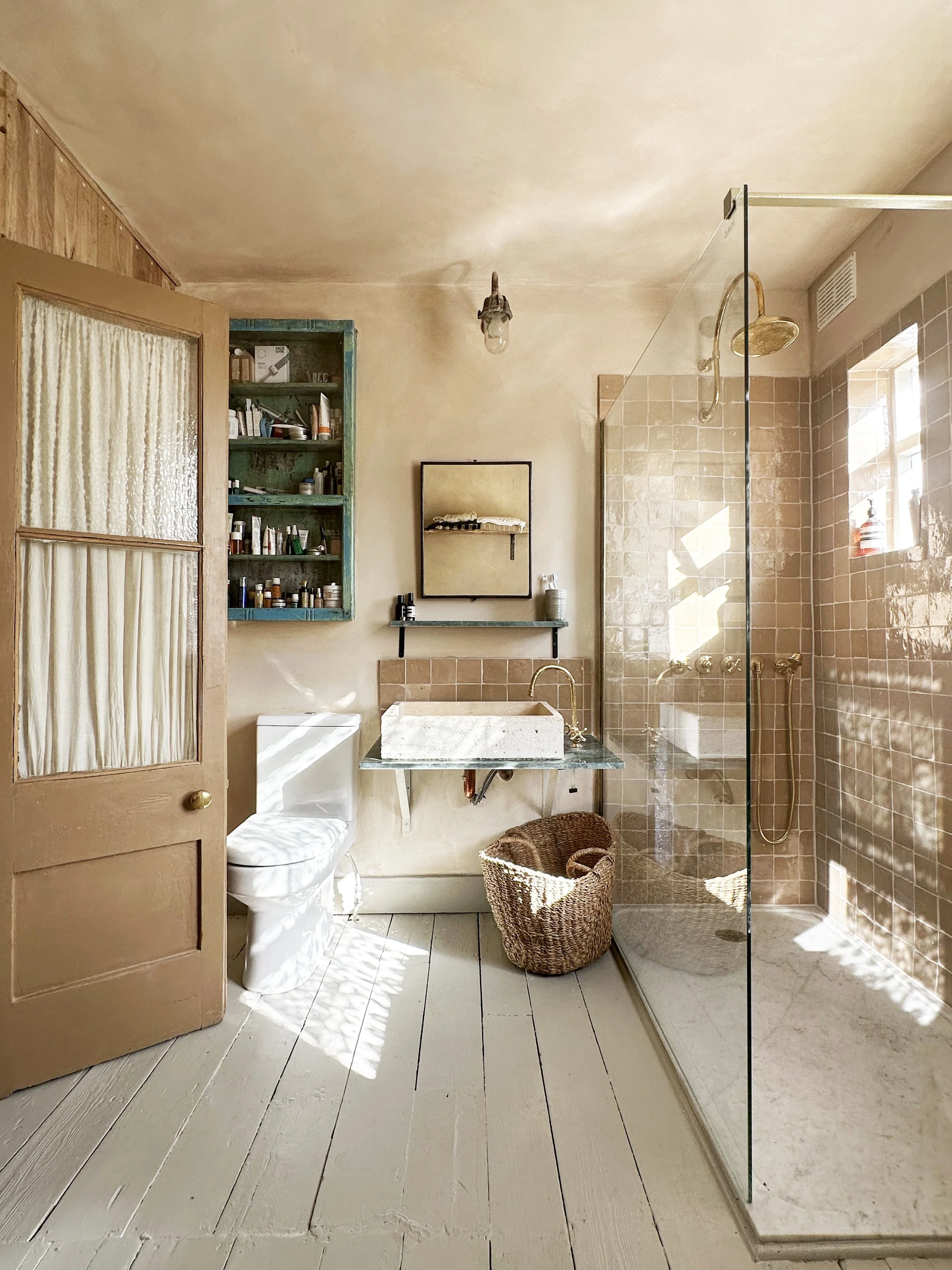 A bathroom with a rustic style featuring a glass shower enclosure, a woven basket, a vintage wooden door with glass panels, a toilet, a concrete sink with a gold faucet, a mirrored cabinet, a small shelf with toiletries, wooden shelves with bottles, 