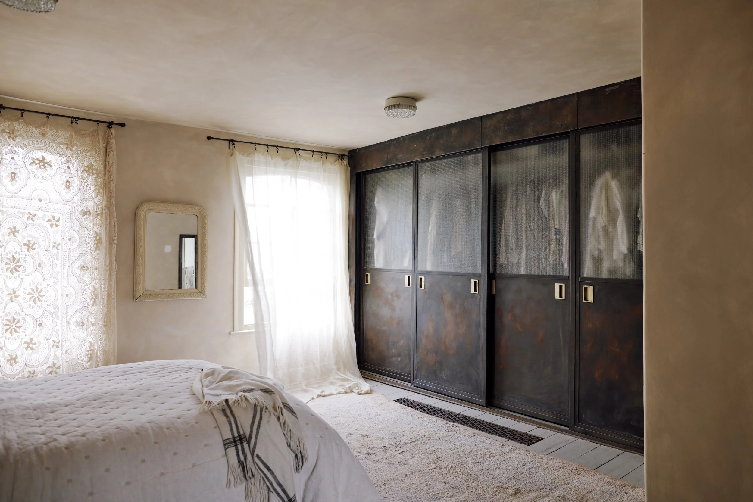 A bedroom with cream-colored curtains, a bed with a white quilt, a wall mirror, a window, and a large black closet with sliding doors.