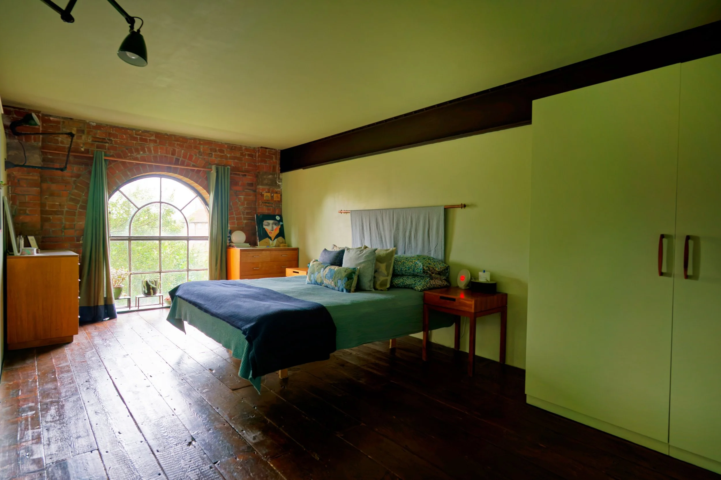 A bedroom with a large arched window, wooden flooring, green walls, and a bed with pillows. There is a small nightstand with a red alarm clock and tissue box, a dresser near the window, and a painting on top of it. The ceiling is yellow with a black 