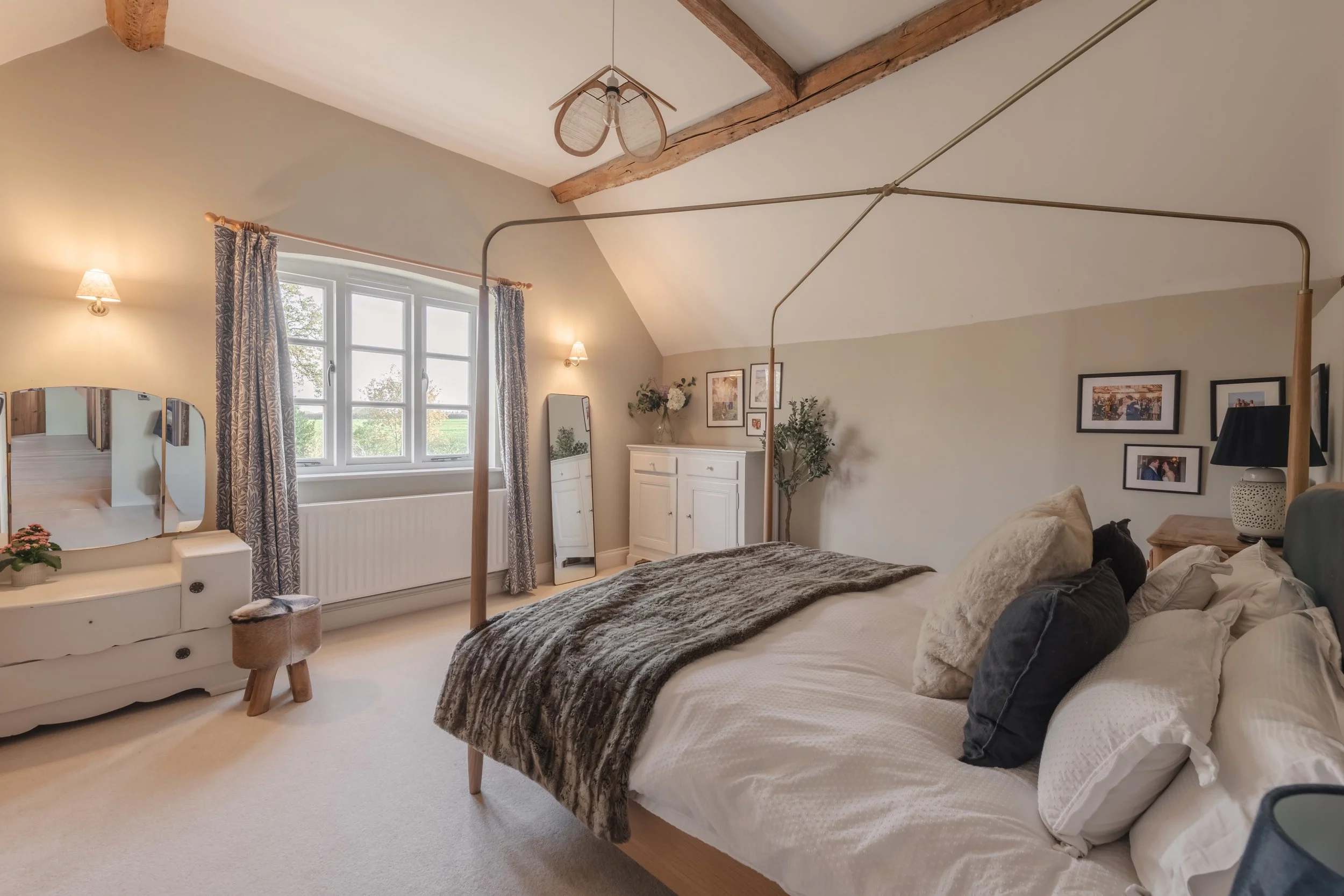 A cozy bedroom with a large bed, decorative pillows, and a throw blanket. A window with patterned curtains, a white dresser with a mirror, a small stool, a cabinet, and framed pictures on the wall. A ceiling light fixture and a lamp on a bedside tabl