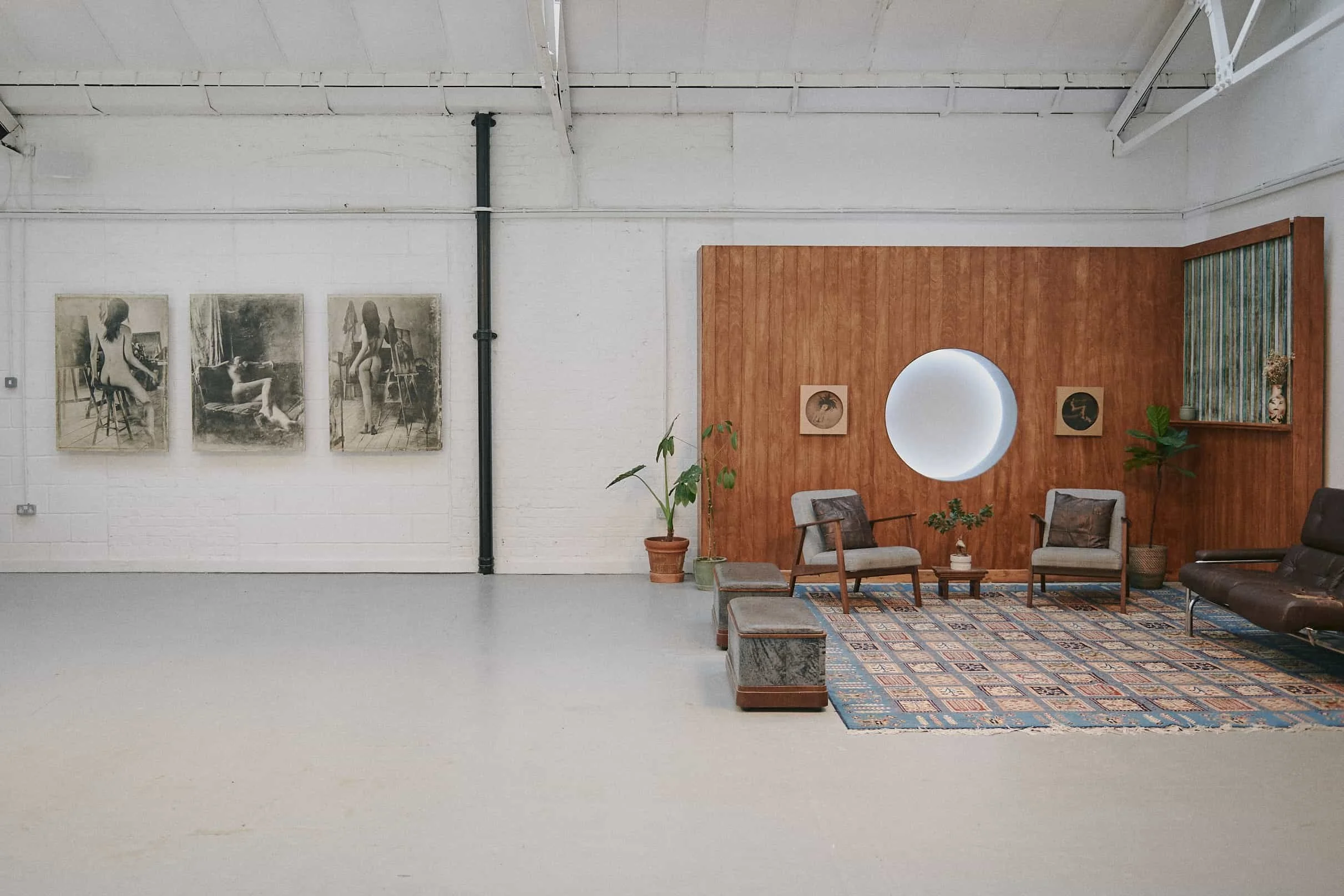 Minimalist waiting area with vintage-style armchairs, a patterned area rug, and potted plants, against a white brick wall with framed artwork and a wooden accent wall with a circular window.