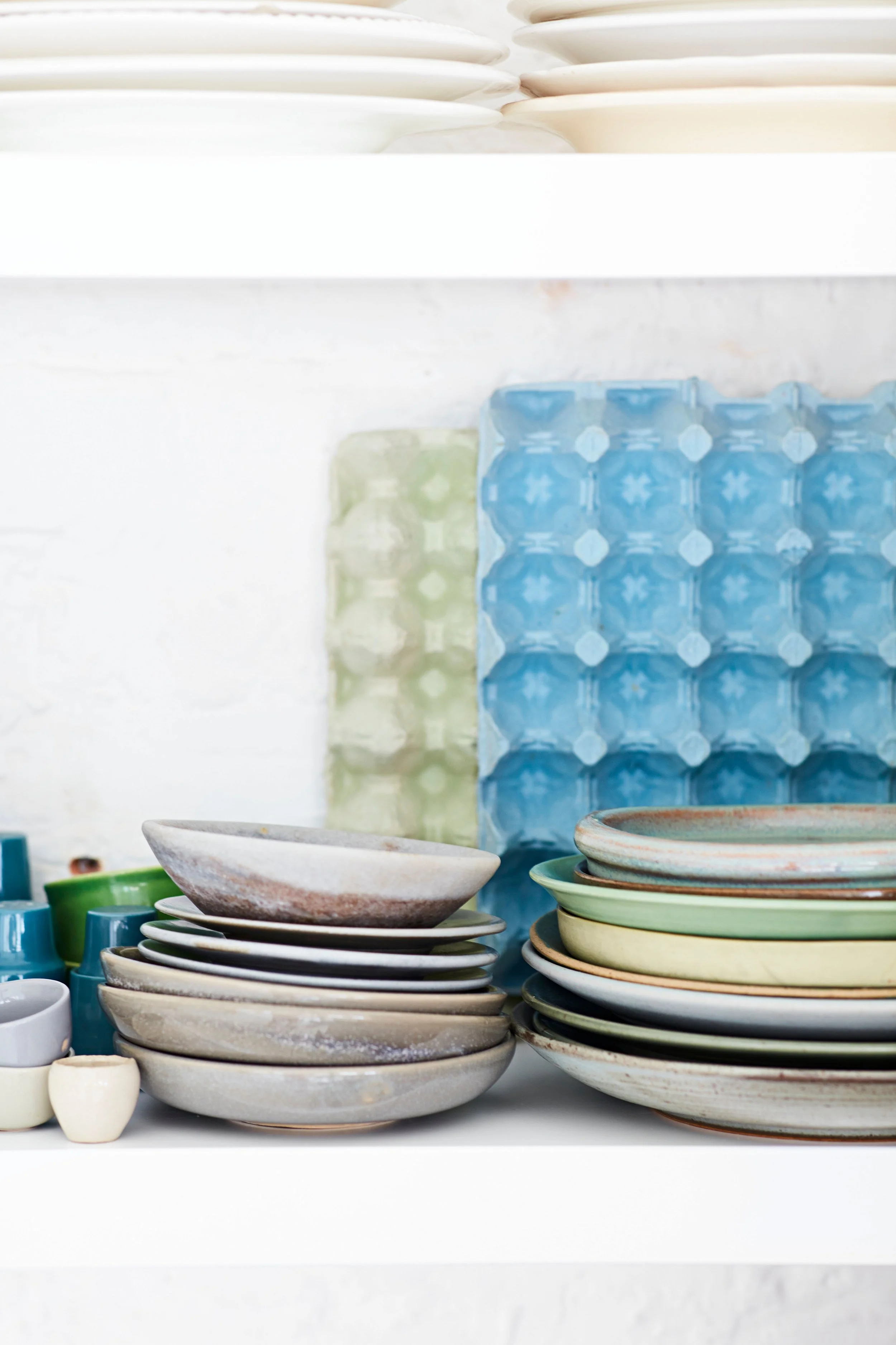 Stacks of ceramic bowls and plates in various colors on a white shelf with a white brick wall background.