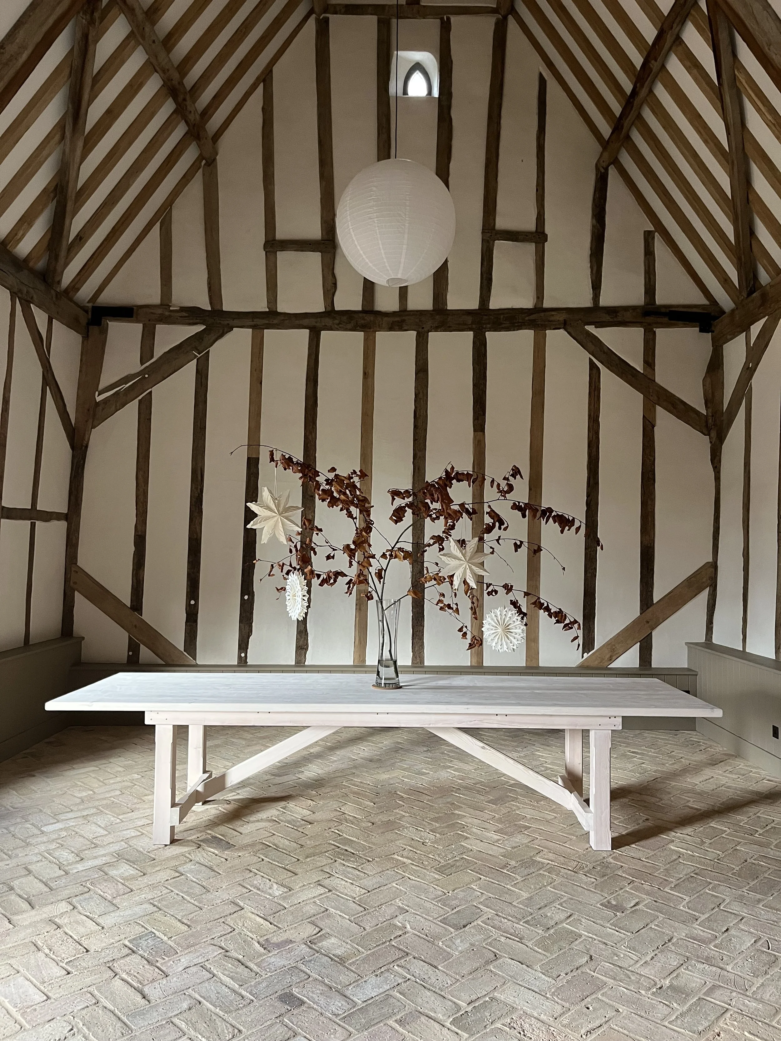 A white wooden table with a glass vase holding a branch with brown leaves and floating paper decorations, set in a rustic room with exposed wood beams and a brick floor, decorated with a paper lantern and hanging paper snowflakes.