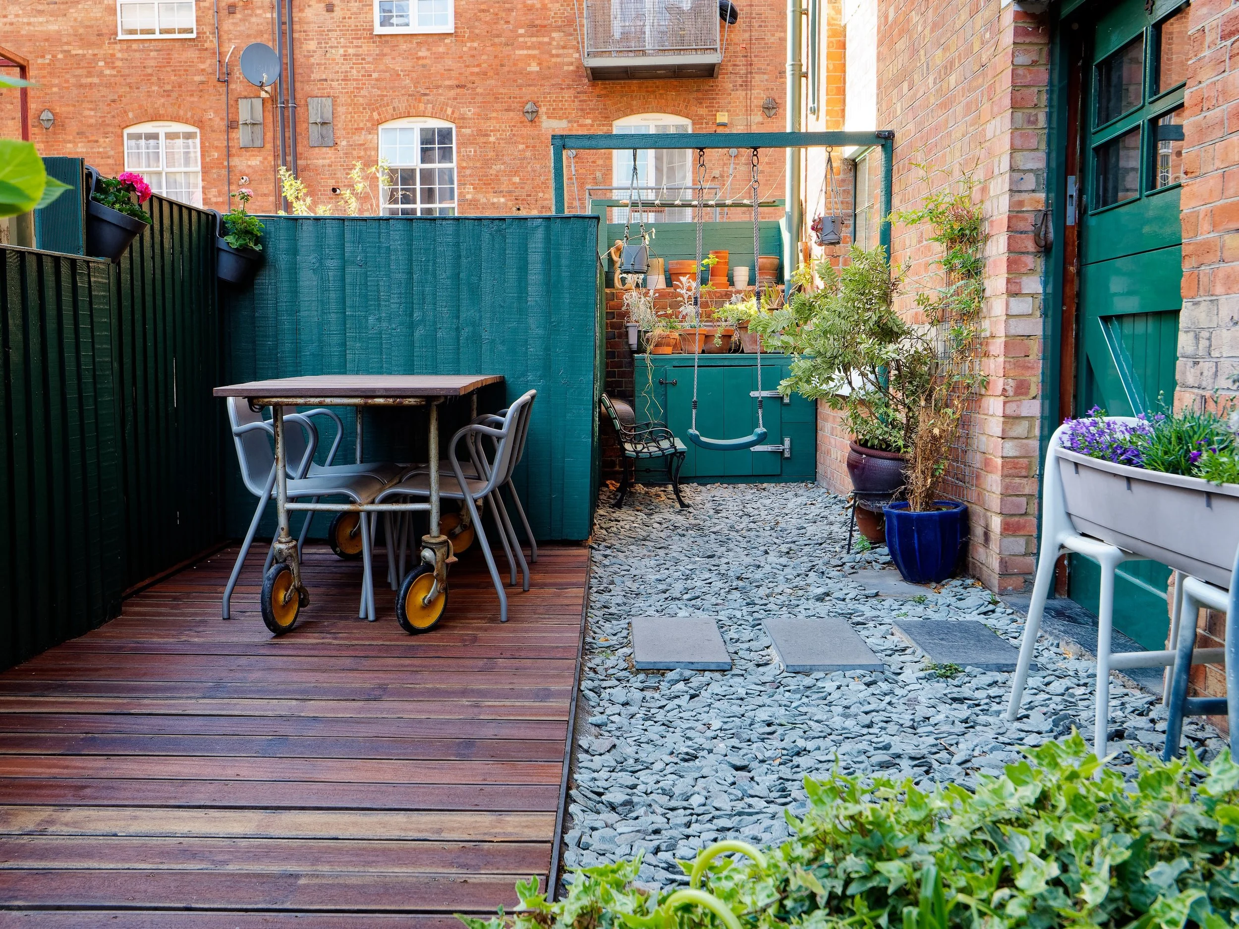 Small outdoor patio with wooden decking and a gravel pathway, featuring potted plants, a table with stacked chairs, and a green gate.