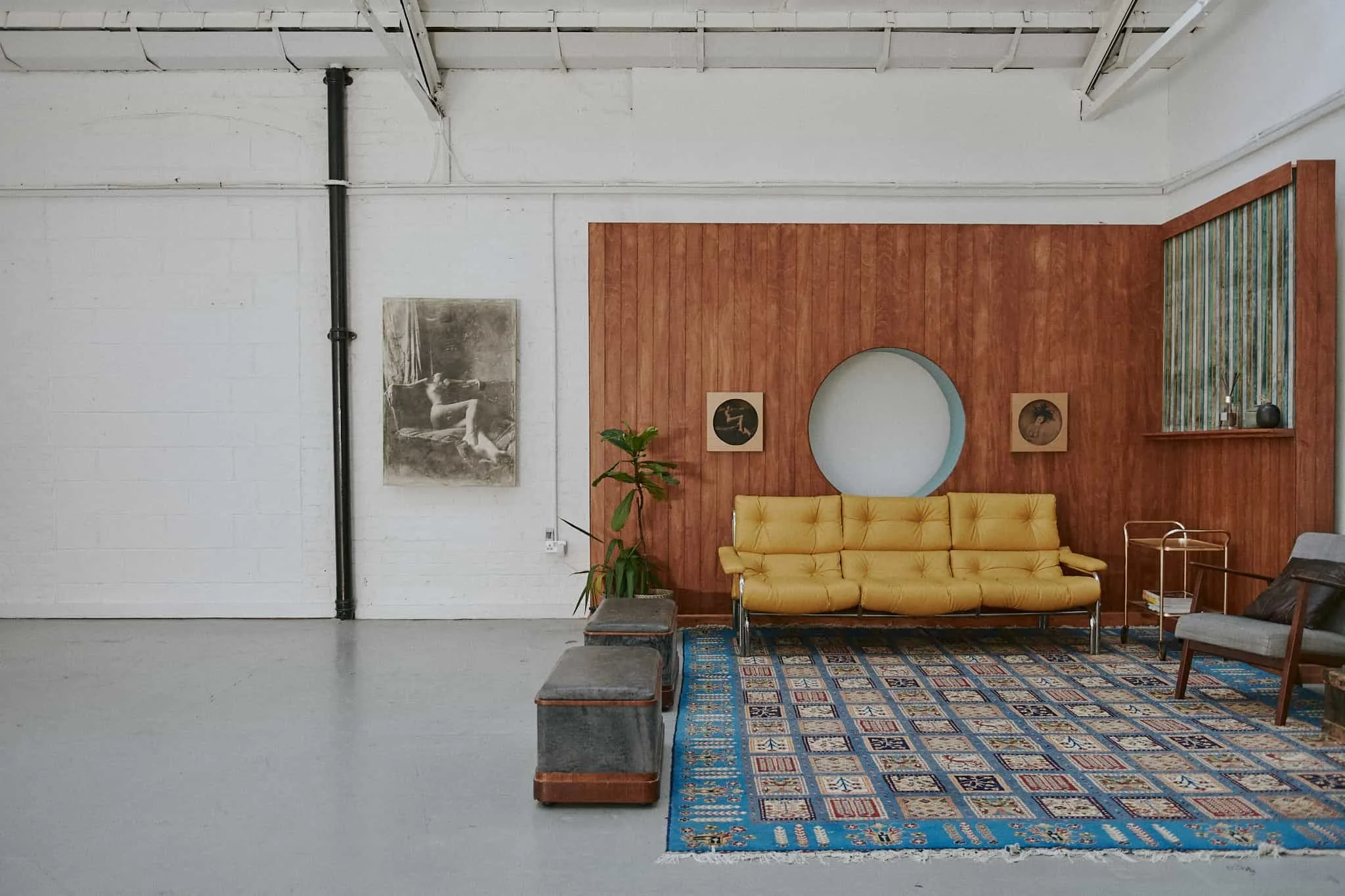 Modern living room with white brick walls, a wood-paneled accent wall, and a yellow sofa on a patterned blue rug. There is a potted plant, framed artwork, and a small side table with decor.