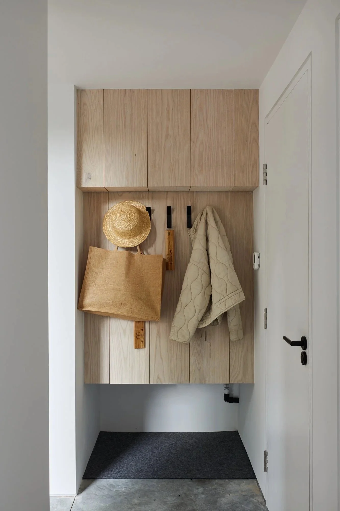 Entryway with a wooden wall-mounted coat rack holding a straw hat, a tote bag, and a quilted jacket. Above the rack there are wood cabinets. To the right is a white door with a black handle.