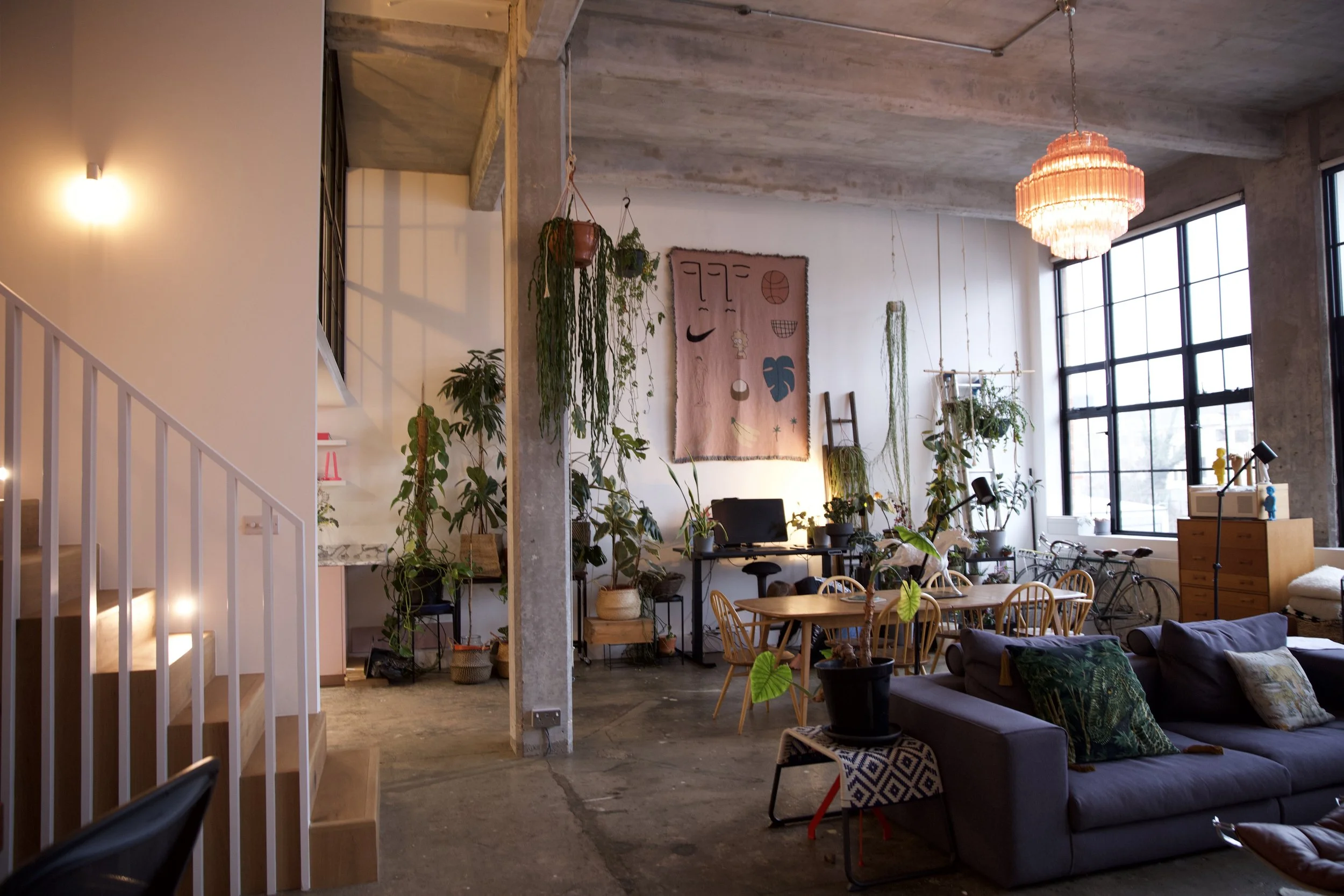 Interior of a loft-style living space with large windows, concrete floors, a gray sofa with cushions, a wooden dining table with chairs, numerous hanging and potted plants, a colorful chandelier, and art on the wall.