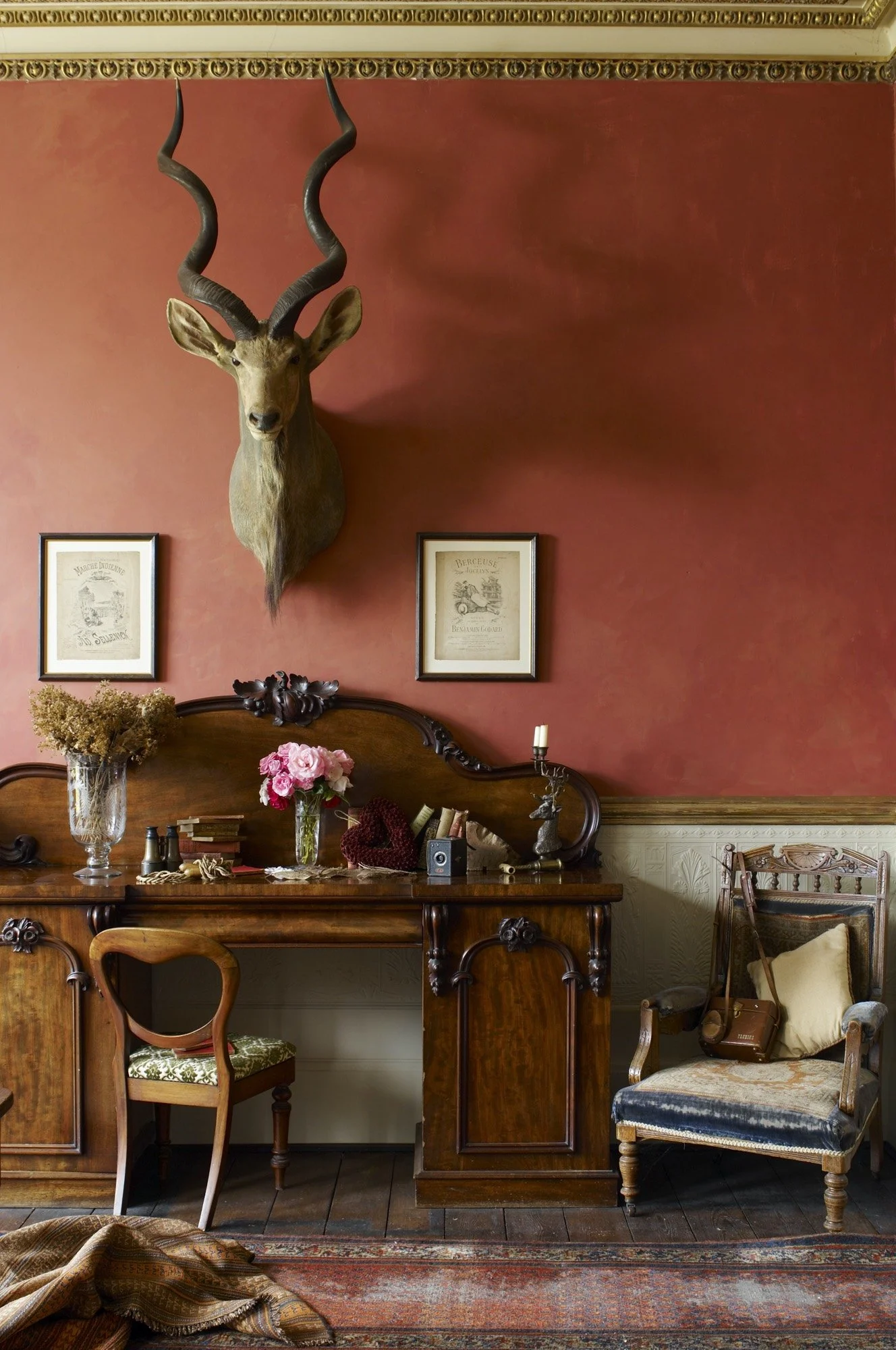 A vintage interior with a mounted deer head on a red wall, two framed sketches, a wooden desk with flowers and various items, a wooden chair, and an antique armchair with a bag and pillow.