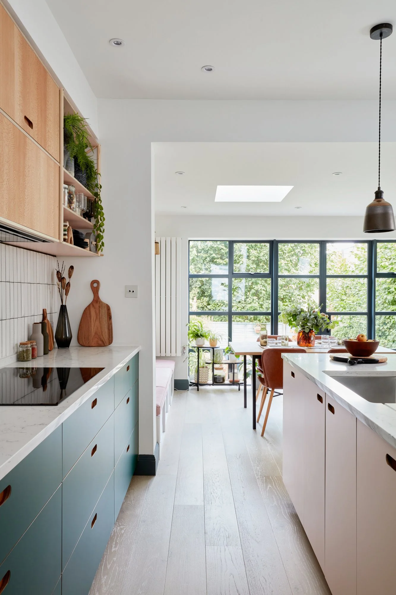 Modern kitchen with light wood flooring, white and teal cabinets, open shelves with plants and dishes, and a dining area with a large window showing green trees outside.