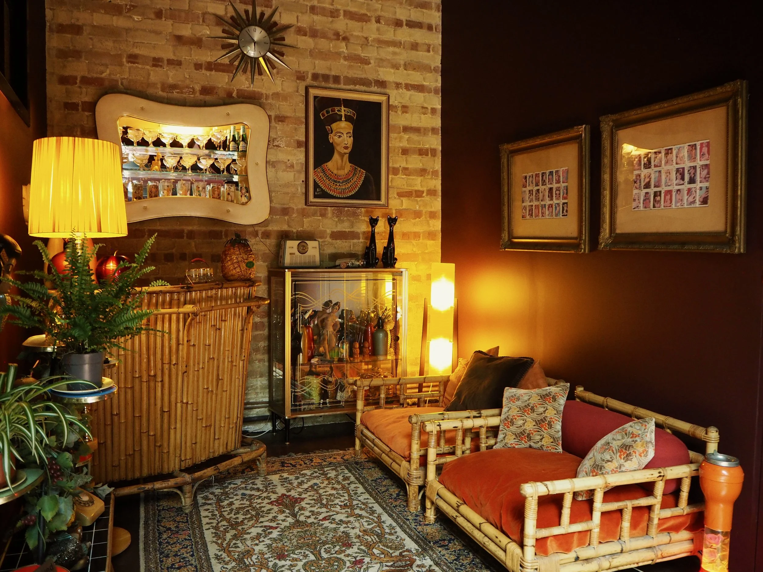 Interior of a cozy living room with a brick wall, bamboo furniture, potted plants, framed art, and warm lighting.