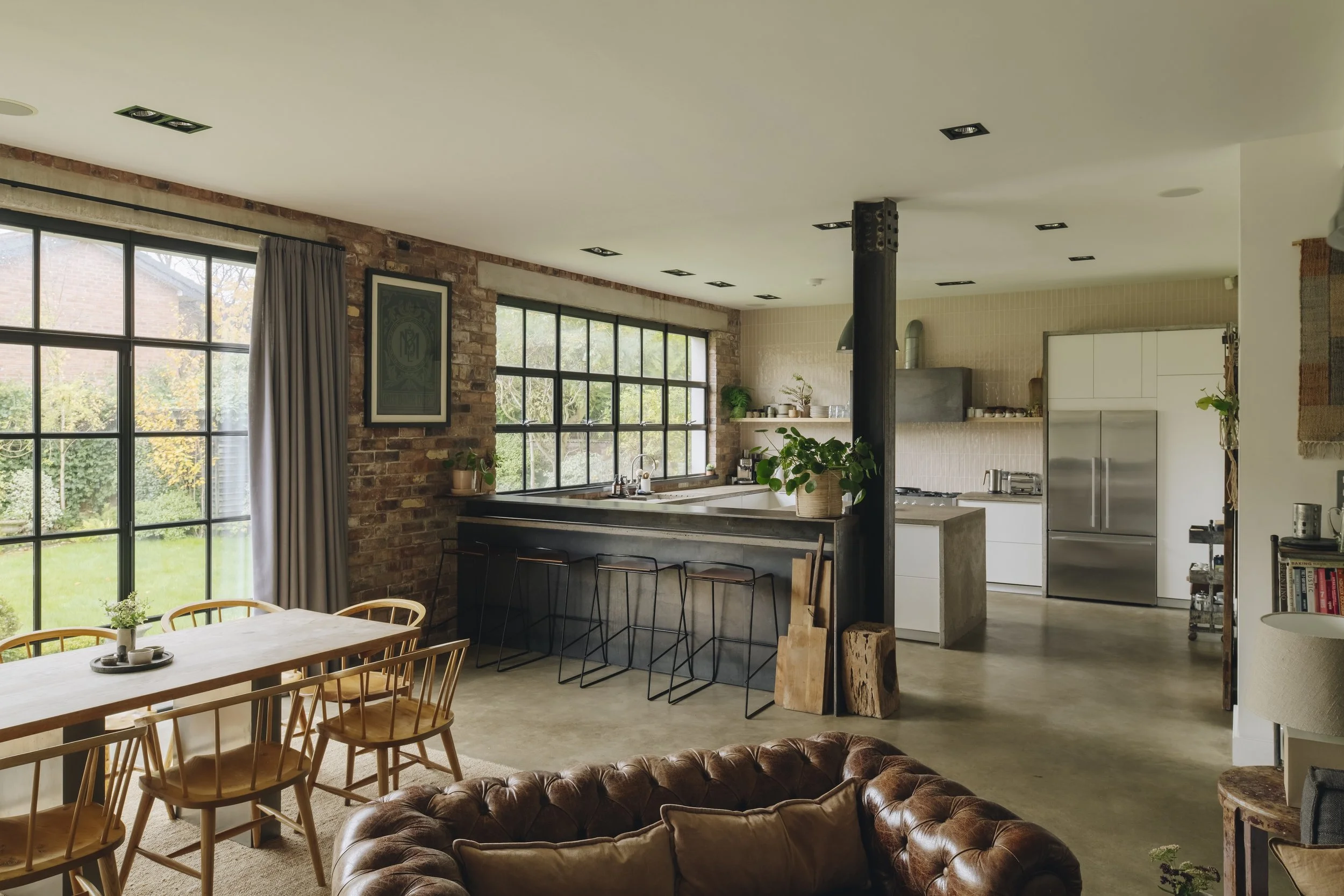 Open-concept living and kitchen area with large windows, brick walls, and modern furniture. A dining table with wooden chairs is near the window, and the kitchen features white cabinets, a stainless-steel fridge, and a counter with stools. There are 