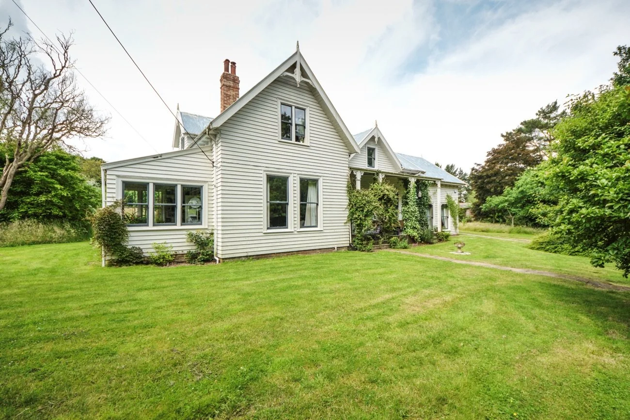 White historic house with decorative trim, surrounded by lush green lawn and trees, under a partly cloudy sky.