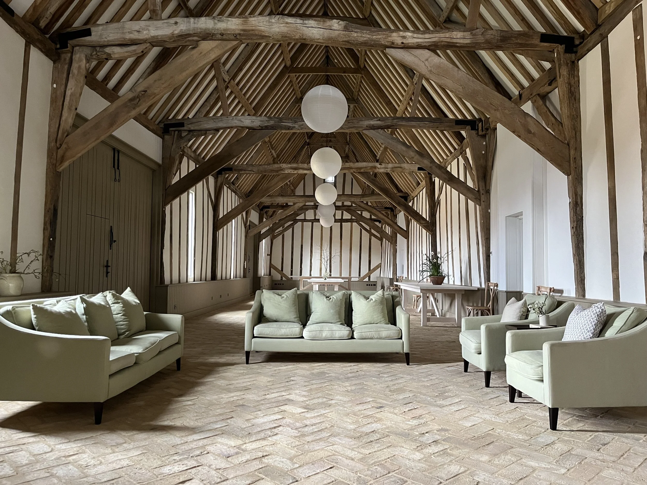 A spacious room with a vaulted wooden ceiling, large wooden beams, and hanging round white paper lantern lights. The room is furnished with light green sofas, a small garden table, and chairs, and has a light-colored brick or tile floor.