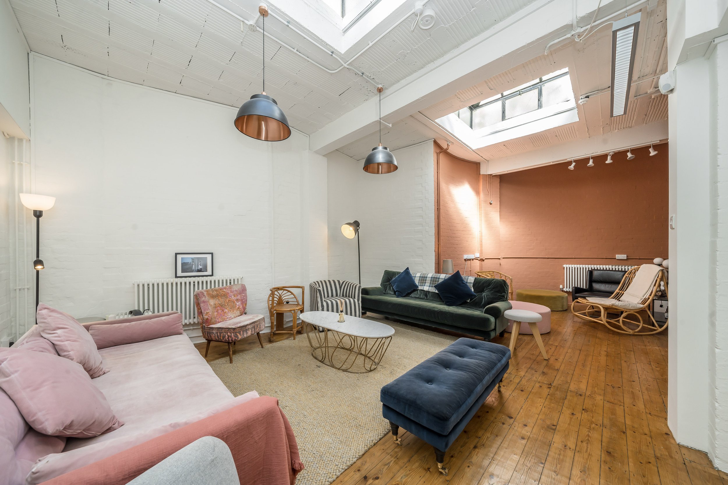 Living room with white brick walls, wooden flooring, large skylight, and modern furnished seating including a pink sofa, green sofa, black armchair, wooden rocking chair, and ottomans, decorated with cushions and lamps.