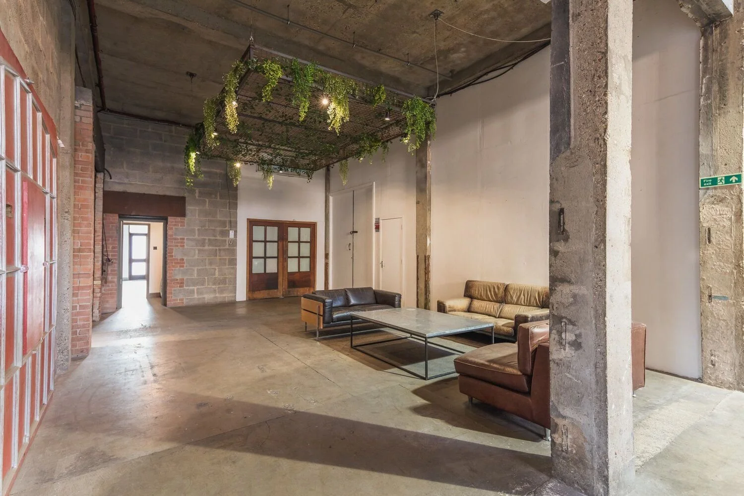 Industrial-style lounge area with leather sofas, a coffee table, exposed brick and concrete walls, and a decorative ceiling with hanging greenery.