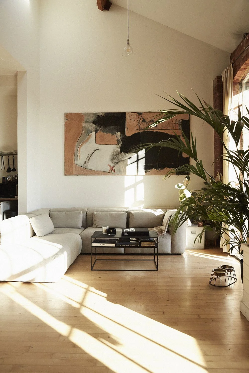 Living room with a white sectional sofa, a black coffee table, a large abstract painting on the wall, a potted plant in the corner, and sunlight streaming through a window, casting shadows on the wooden floor.