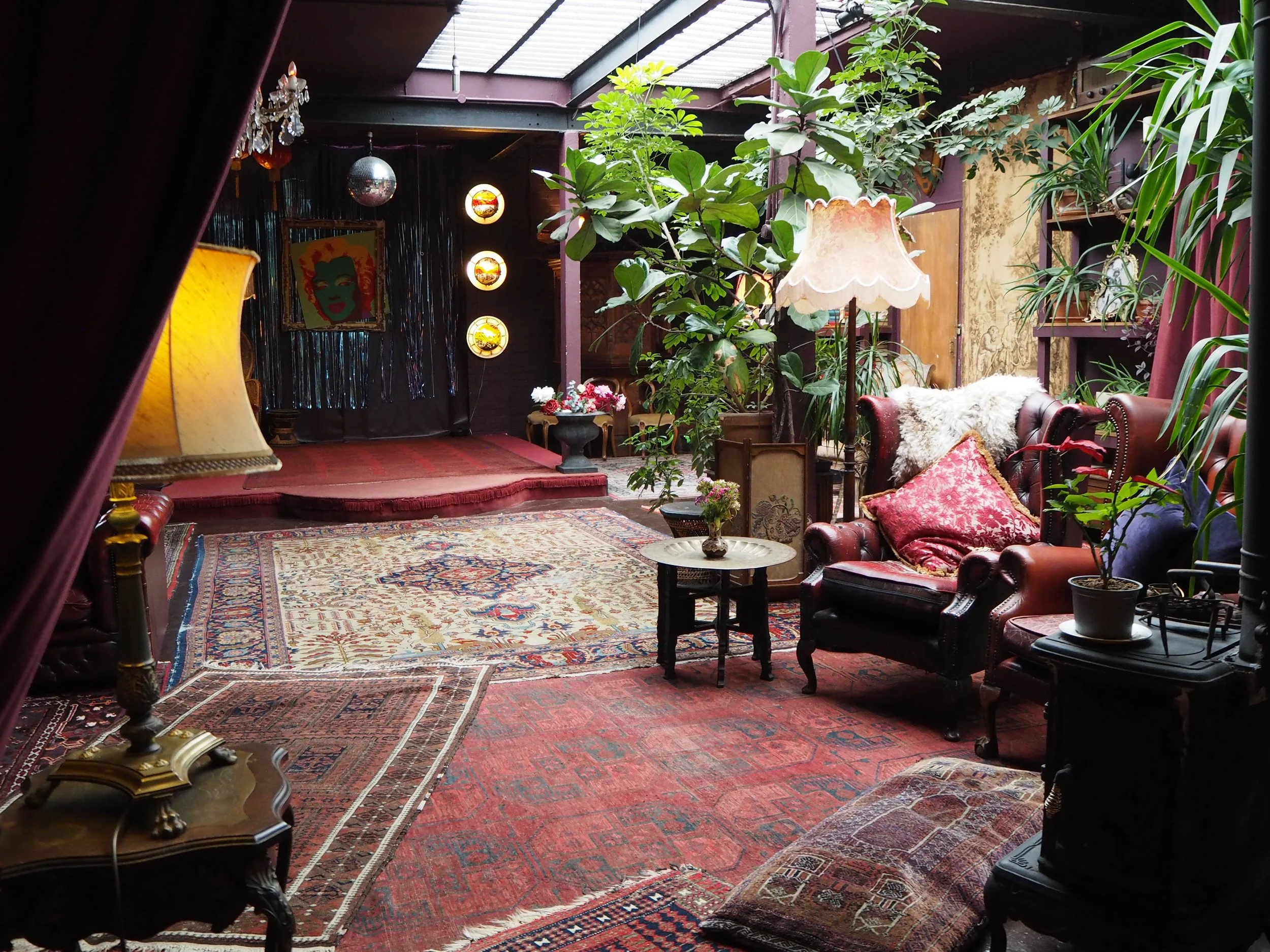 Vintage living room with leather armchairs, Persian rugs, tall indoor plants, and decorative lamps, set on a red carpet with a small table. There is a stage with a curtain, a disco ball, and pop art in the background.
