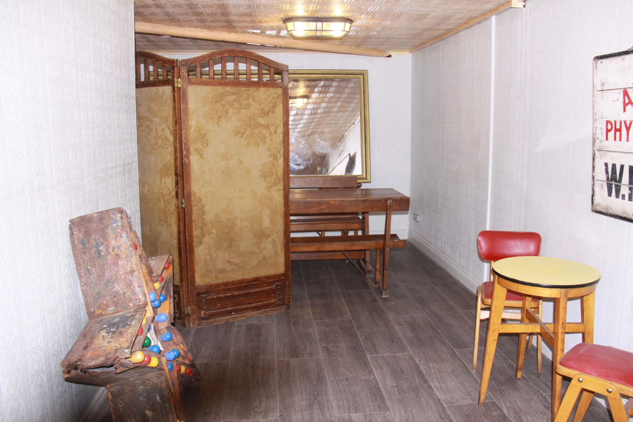 A room with rustic furniture, including a vintage wooden folding screen, a wooden table with benches, a rusty pinball machine, a framed mirror on the wall, a small round yellow table with a red chair, and a wall sign with partially visible text.