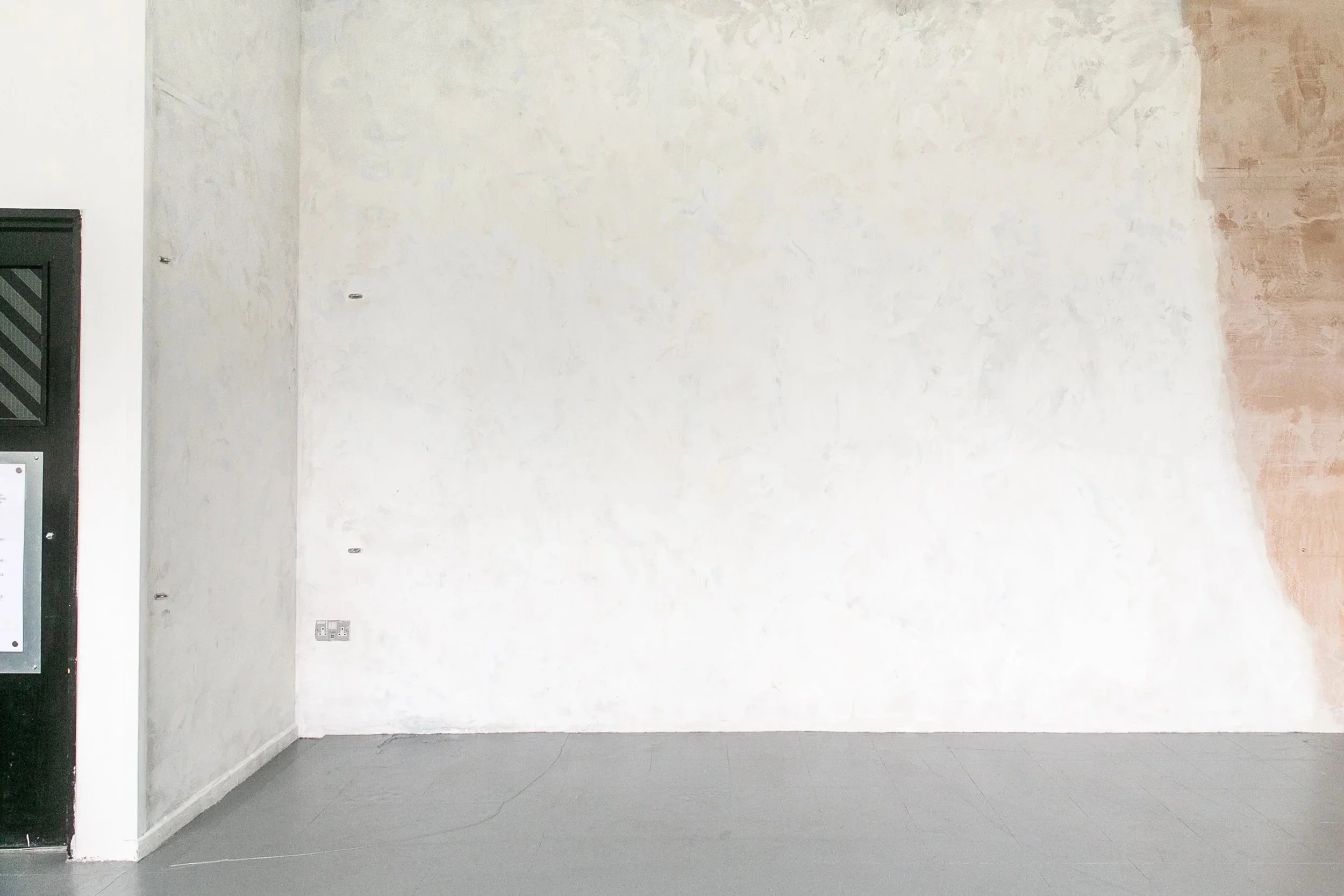 Empty interior wall with some patches of exposed plaster and electrical outlets at the bottom, adjacent to a black elevator door on the left.