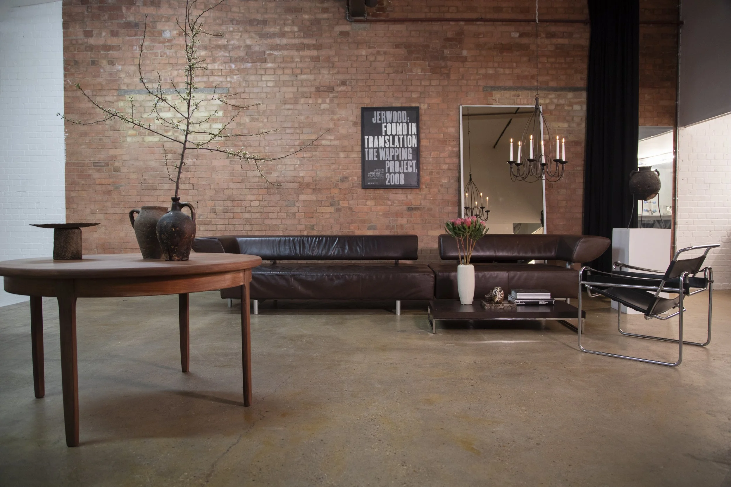 Living room with exposed brick wall, leather sofa, black chair, round wooden table with vases and branches, wall mirror, wall art, chandelier, and decorative items.
