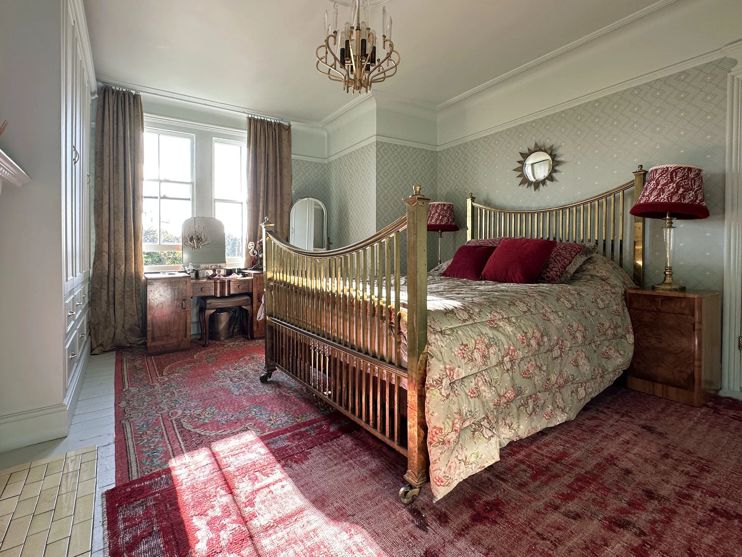 A bedroom with a large bed, gold headboard, floral bedspread, and red pillows, flanked by twin nightstands with red lamps. Sunlight streams in through two large windows with beige curtains. To the left, a vintage vanity with a mirror and various item