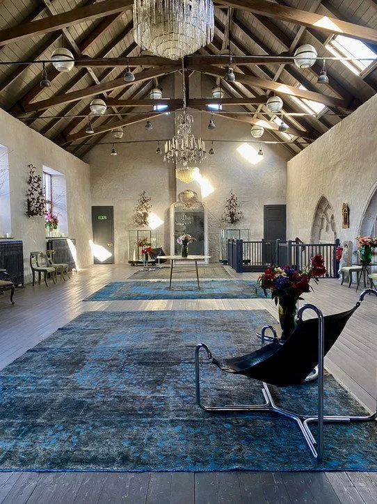 Interior of a church or chapel with high vaulted wooden ceilings, chandeliers, and a blue patterned rug. Decorated with flower arrangements, chairs, and an ornate mirror or altar at the back.