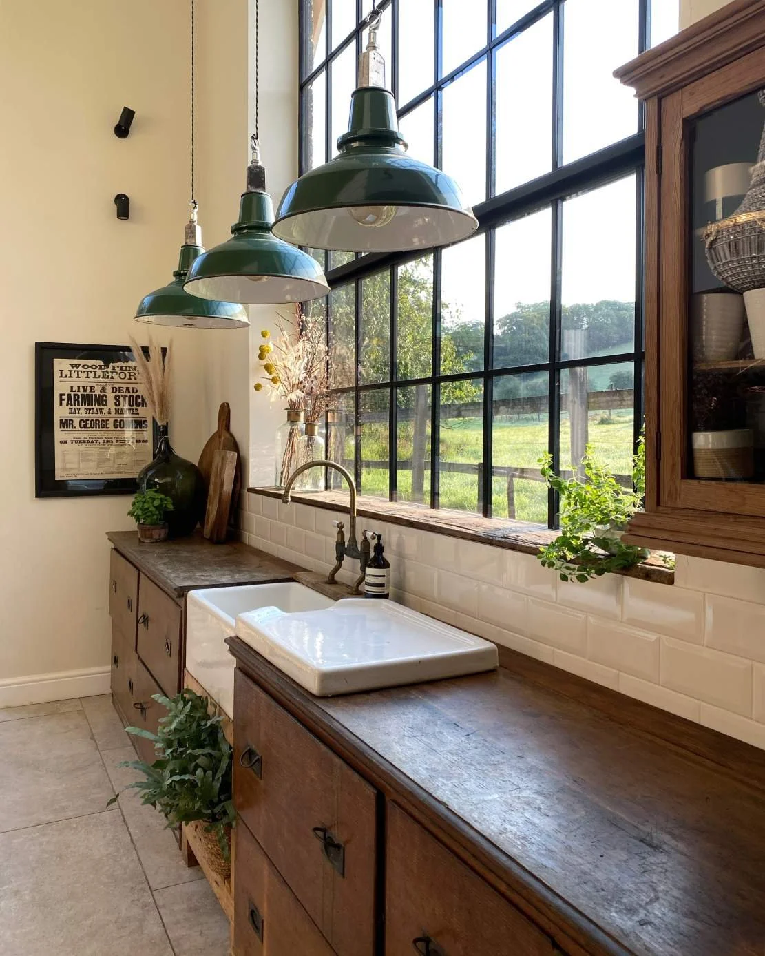 Rustic kitchen with large grid window, farmhouse sink, wooden cabinets, hanging pendant lights, and decorative plants.