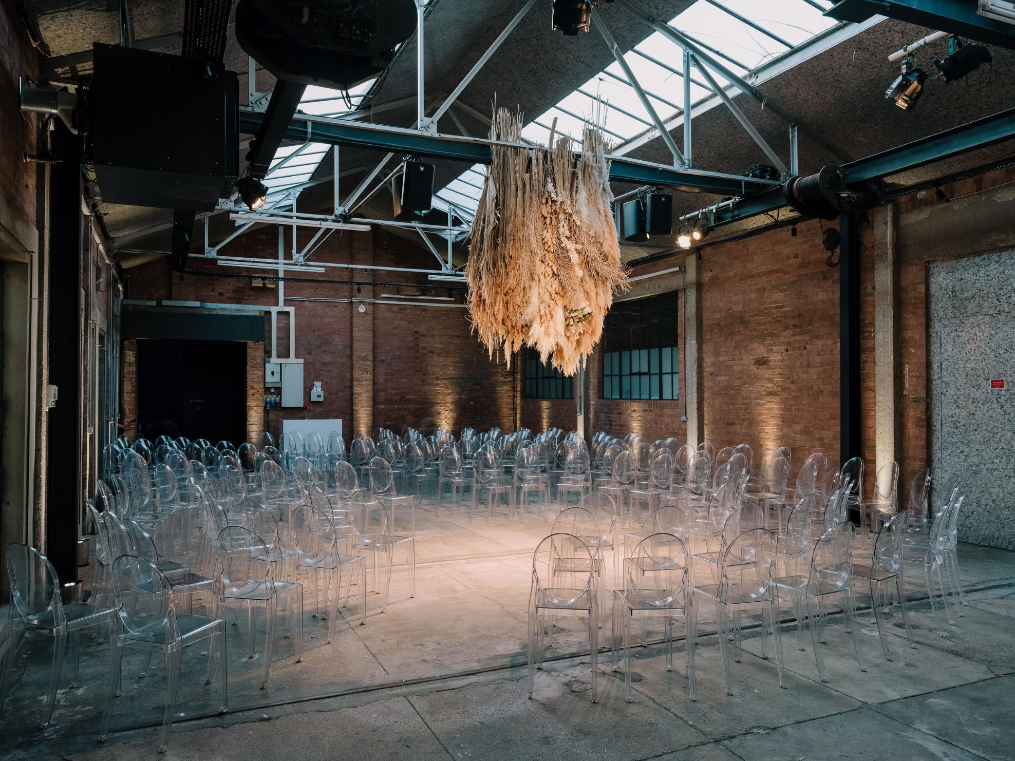 Clear chairs arranged in a circle inside an industrial event space with exposed brick walls and a large hanging decoration made of dried plants.