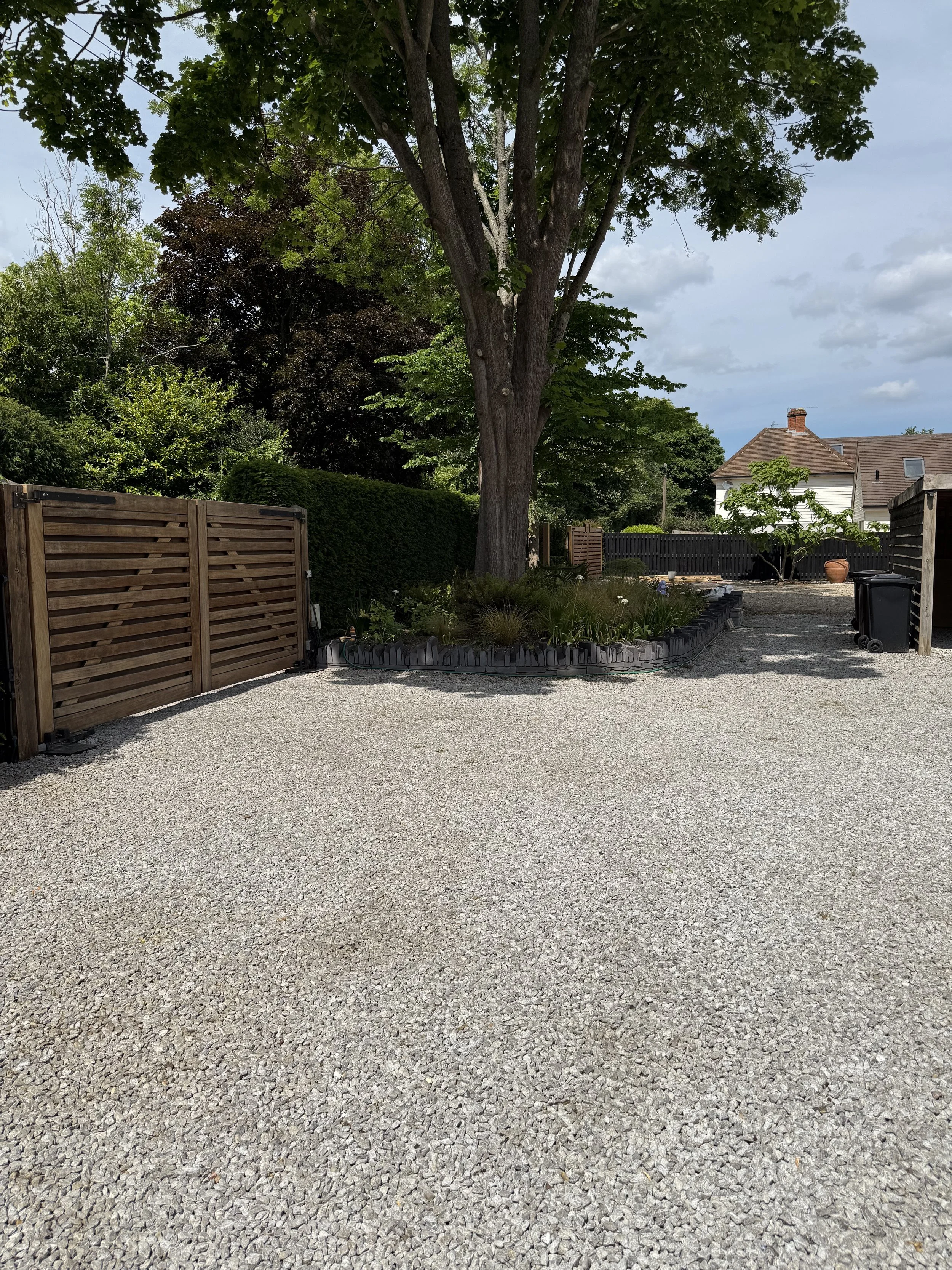 A backyard with a gravel ground, a large tree with green leaves, a flower bed surrounding the tree, and a wooden fence on both sides.