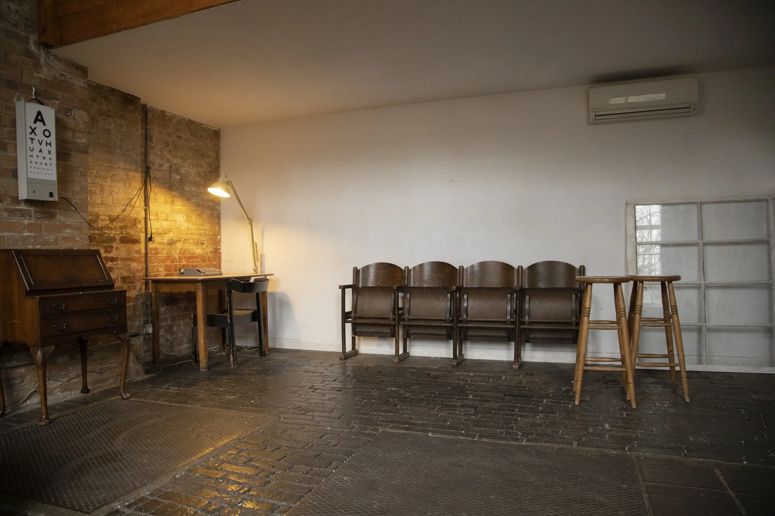 Empty room with brick wall, wooden furniture including chairs and stools, a desk with a lamp, an air conditioning unit, and an eye chart on the wall.