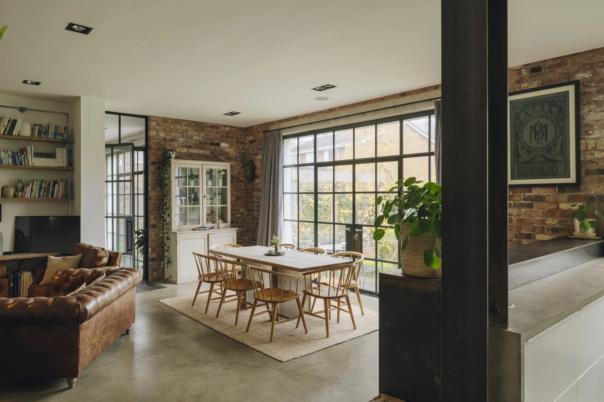 Interior of a modern, rustic-style living and dining room with large windows, brick walls, wooden furniture, and indoor plants.
