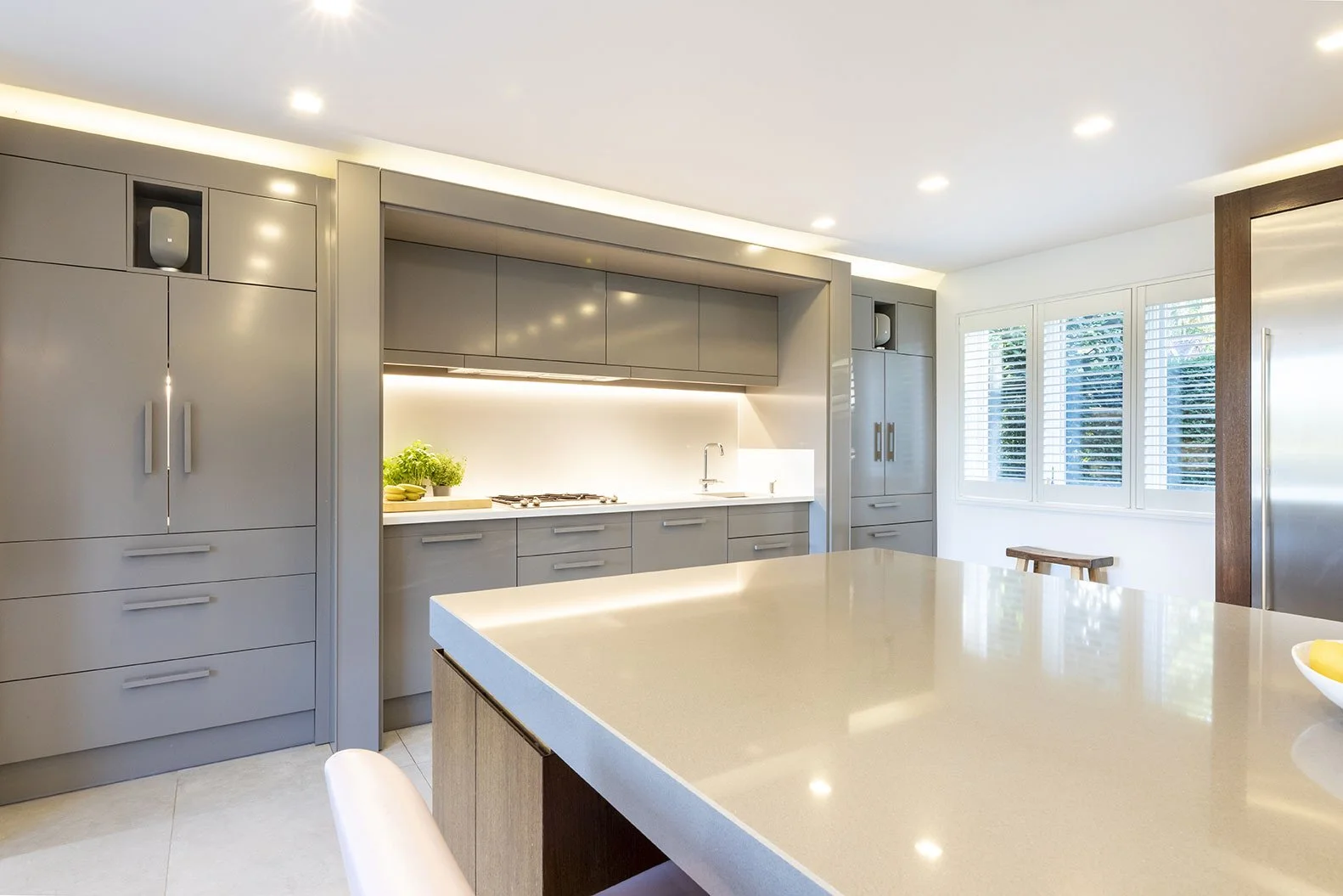 Modern kitchen with gray cabinets, a beige island, white walls, large windows, and recessed ceiling lights.