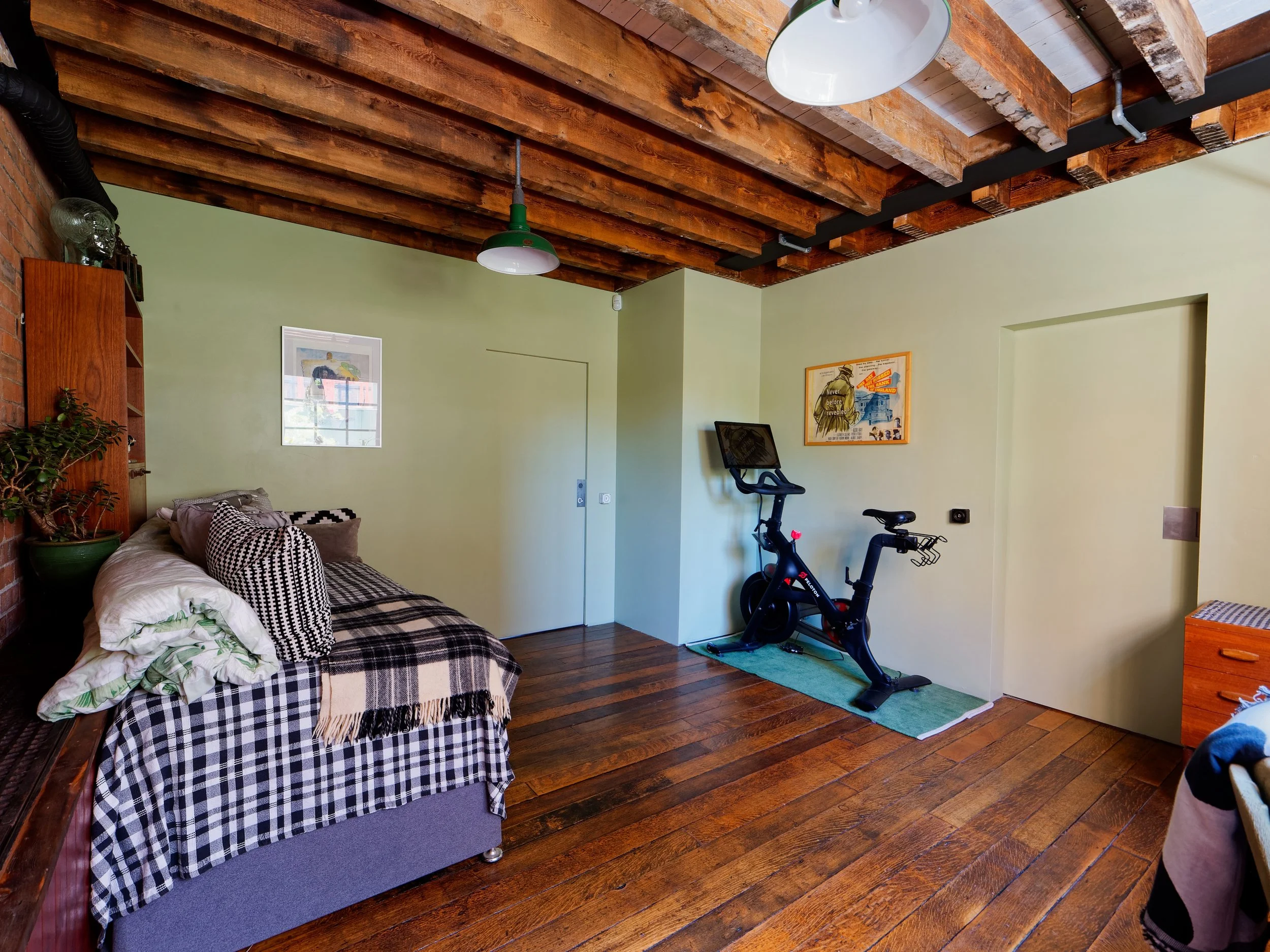 Living room with green walls, wooden ceiling beams, hardwood floor, a bed with black and white checkered bedding, an exercise bike, and framed artwork on the walls.
