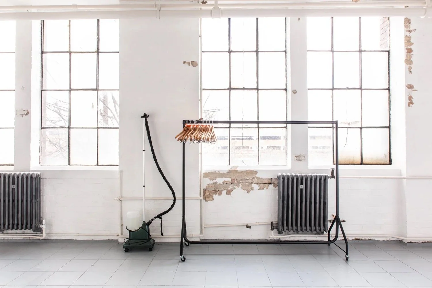 Empty room with large industrial windows, white brick walls, radiators, and an unfinished wall section. Contains clothing rack with hangers and a steamer.
