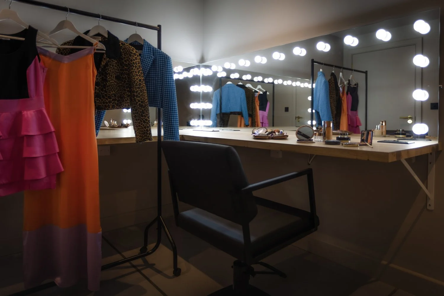 A well-lit dressing room with a large mirror surrounded by Hollywood-style bulbs. There are several colorful dresses hanging on a clothing rack and a vanity table with makeup and accessories.