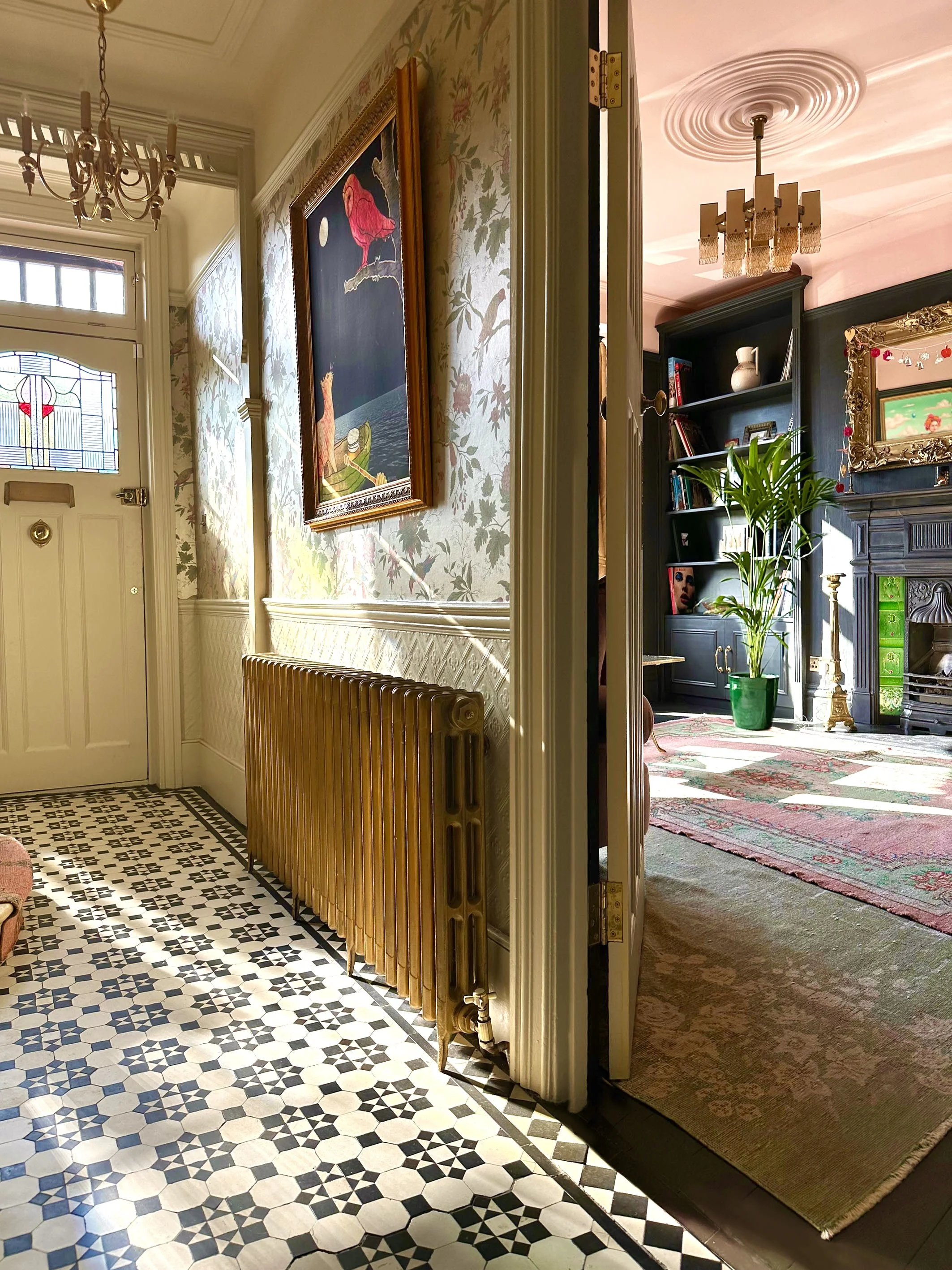 Interior of a house foyer with a black and white geometric tiled floor, a white door with stained glass, wallpapered walls with floral patterns, a chandelier, a framed painting of a pink flamingo, a radiator, and a doorway leading into a living room 