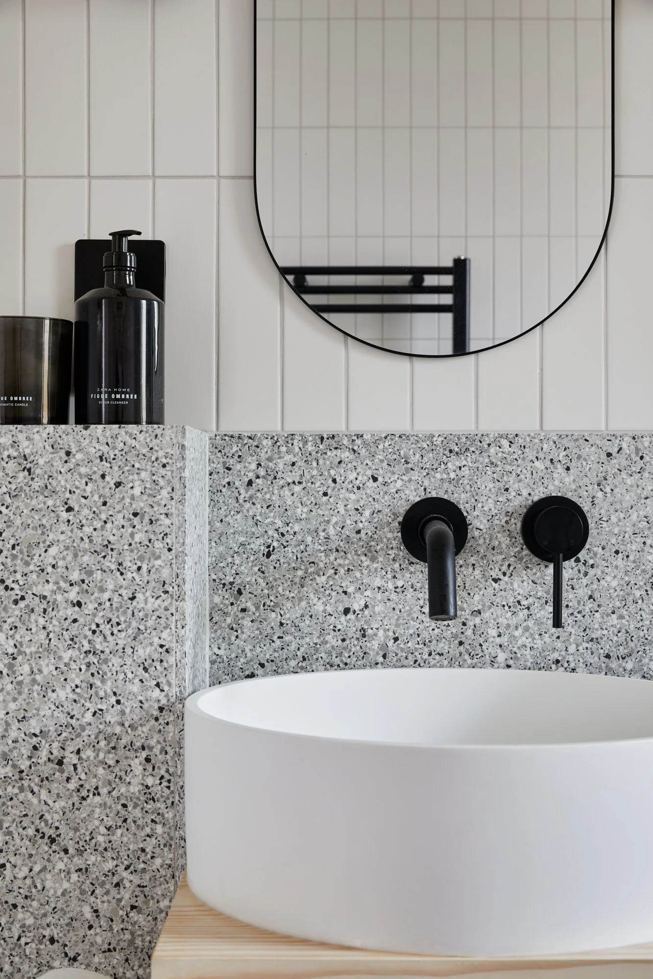 Modern bathroom sink with a white oval basin, black wall-mounted faucet, and a granite countertop. A mirror and toiletries, including black soap dispenser and candle, are visible.